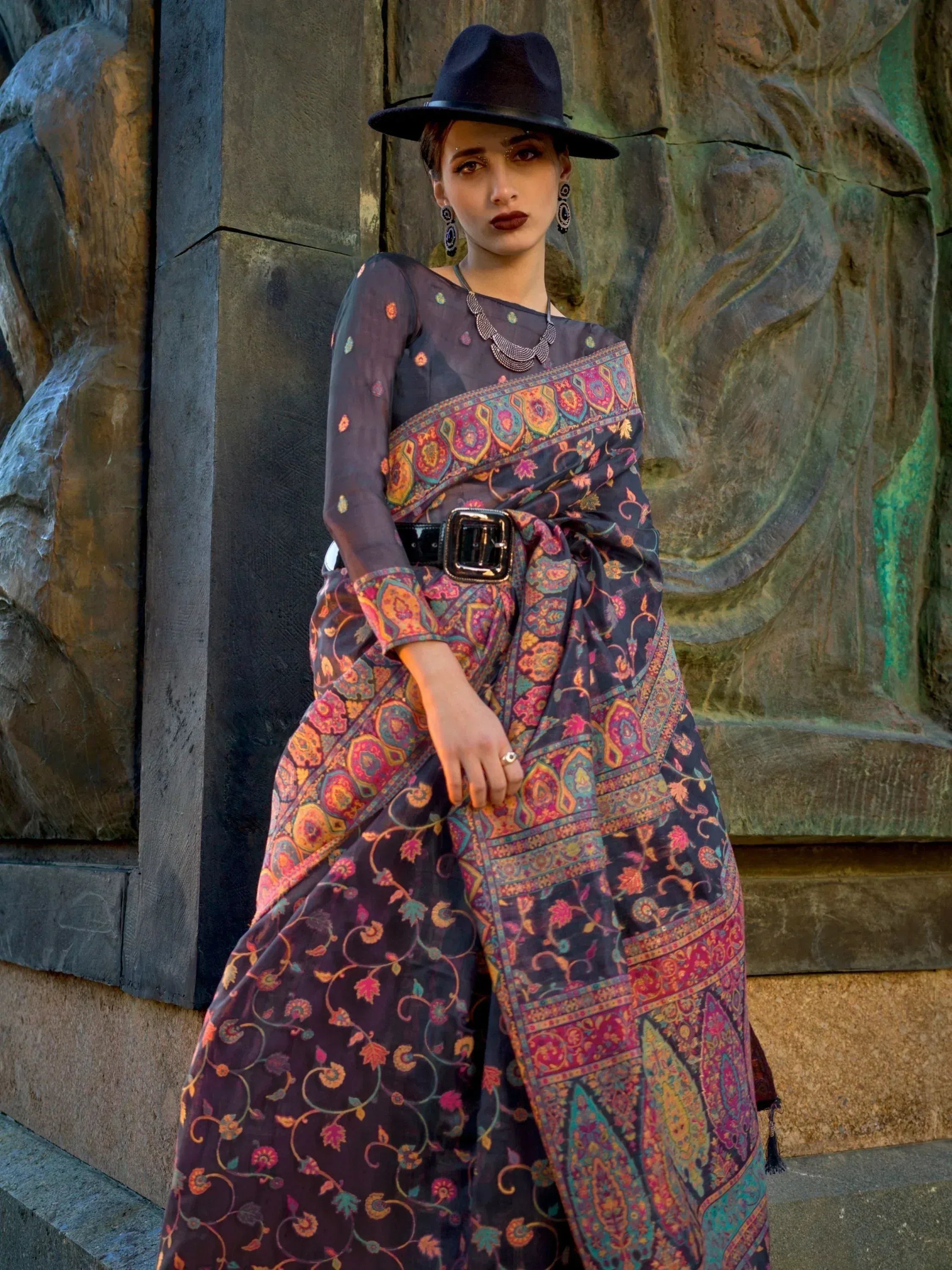 Woman in designer printed saree with belt, black hat, and ethnic jewelry against stone sculpture