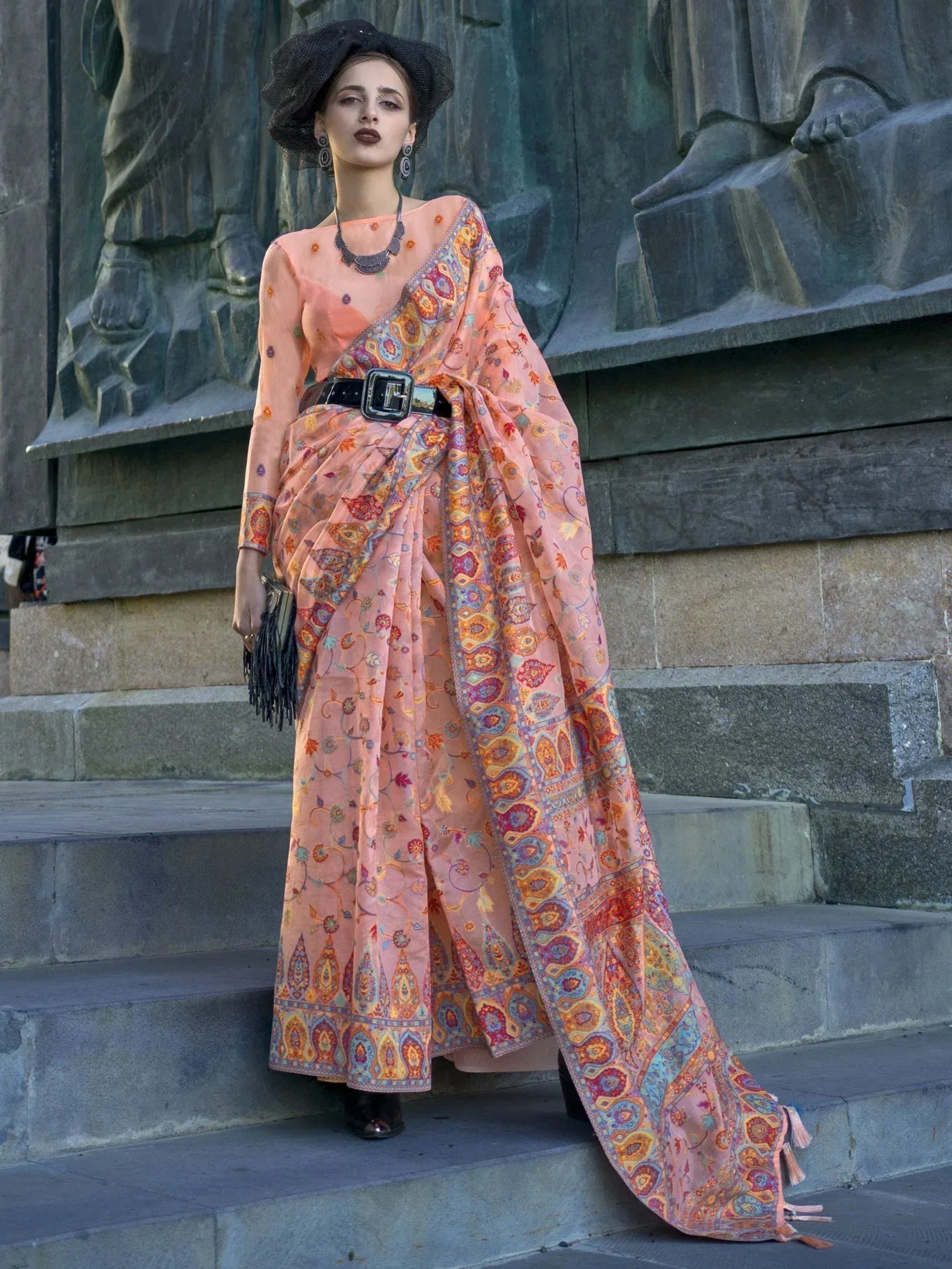 Woman in peach printed saree with belt, black hat, standing on stone steps