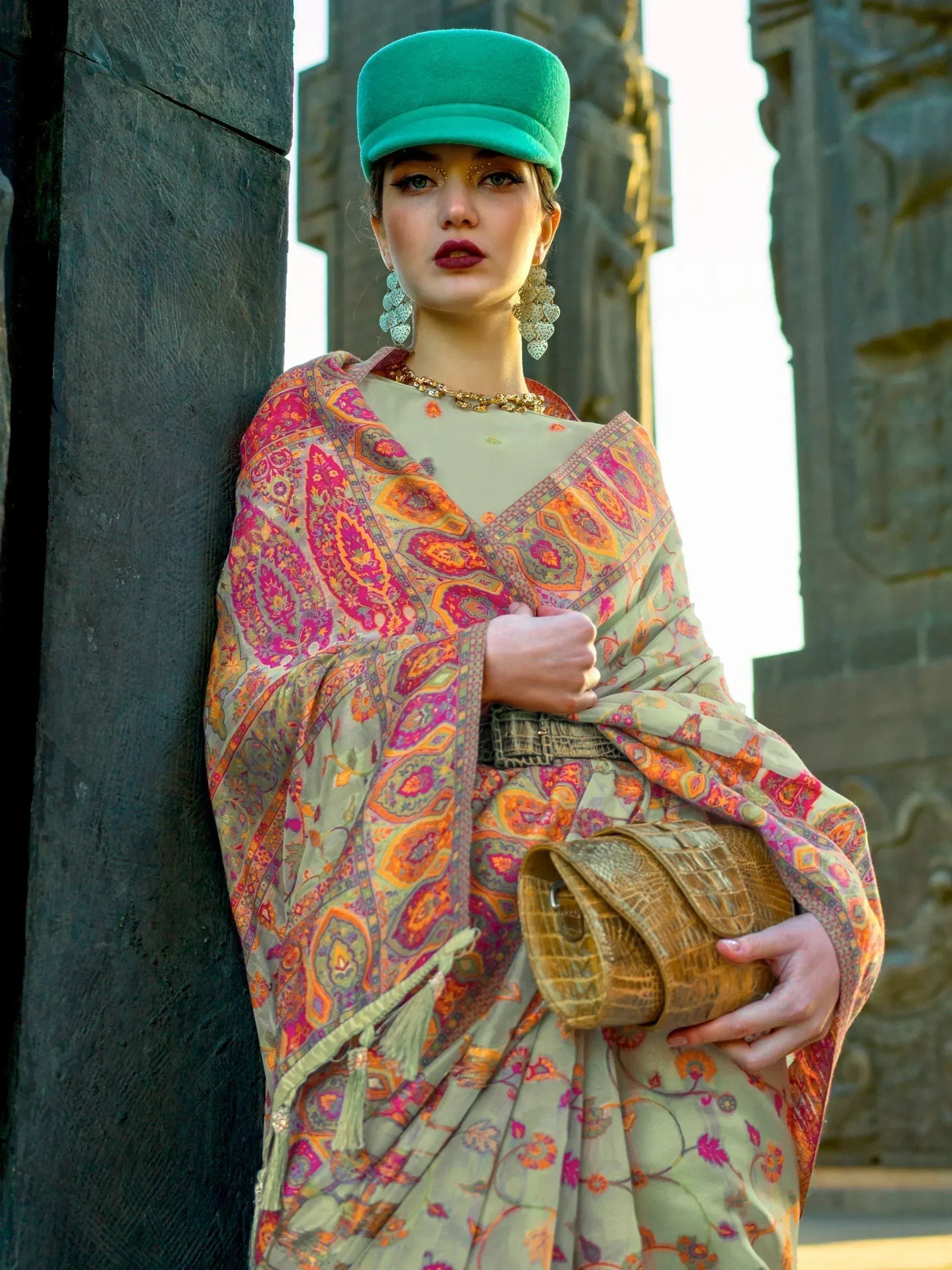 Woman in vibrant ethnic saree with paisley print, teal cap, and gold jewelry, holding textured clutch