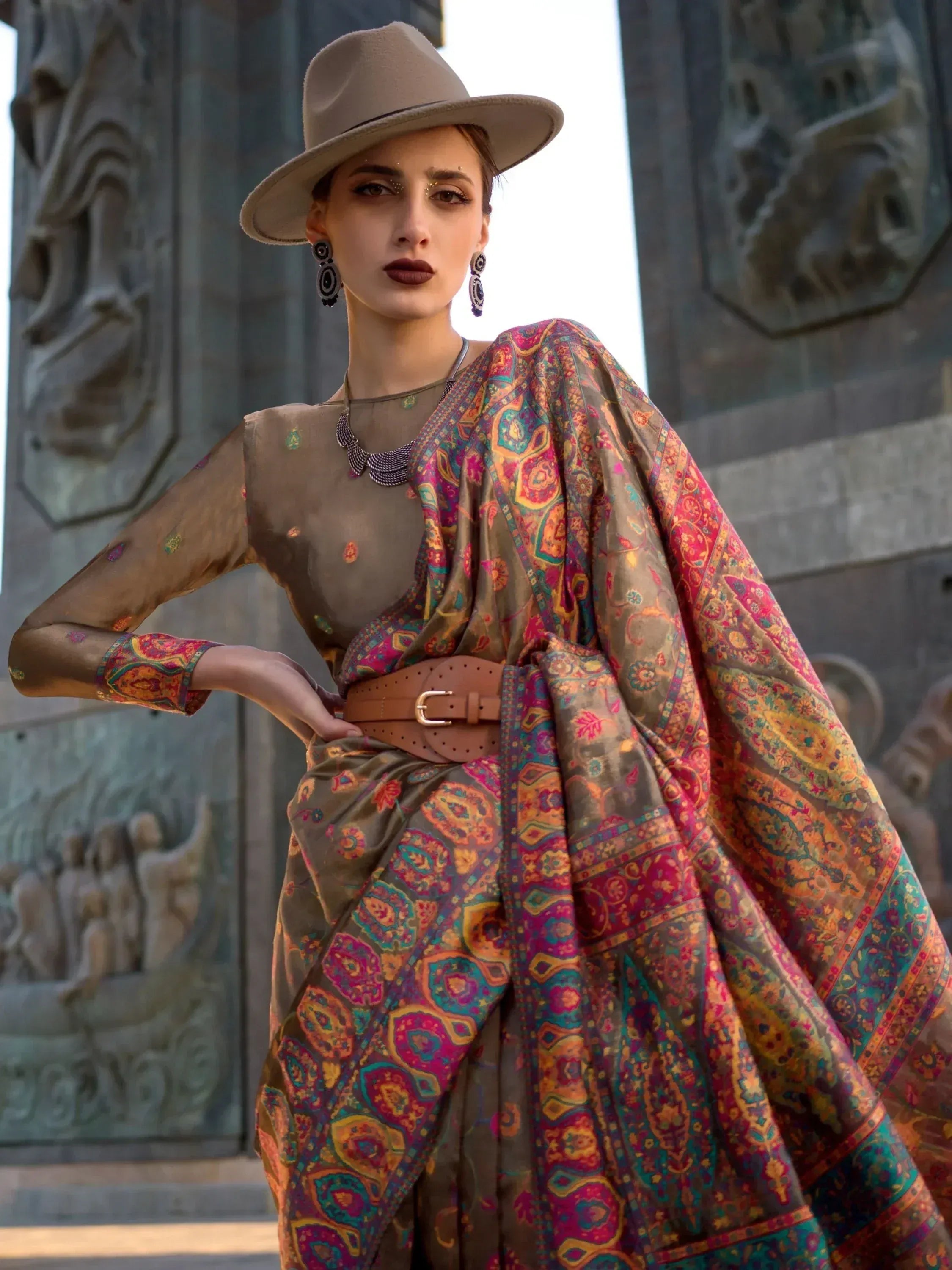 Woman in ethnic printed gown with hat, belt, and statement jewelry, styled for modern Indian fashion.