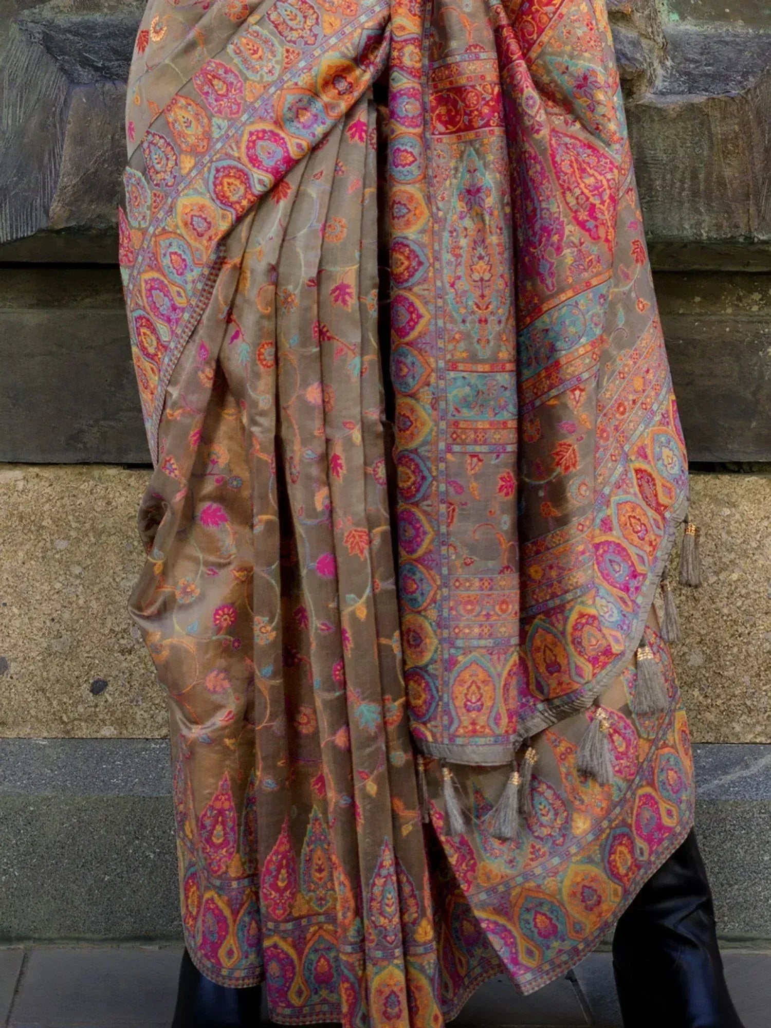Brown ethnic saree with vibrant paisley print and tassels, styled against stone wall