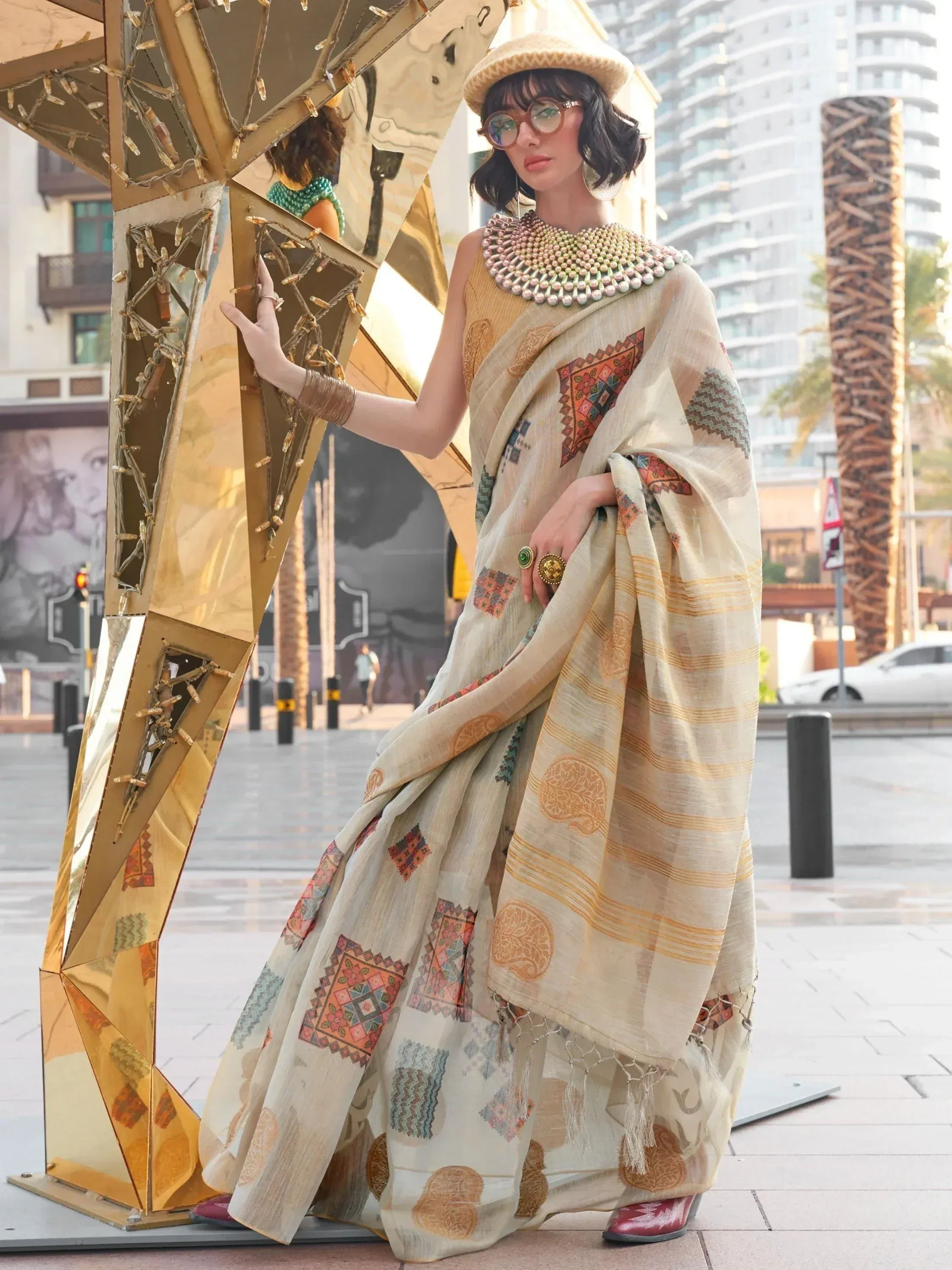 Woman in printed ethnic saree with statement necklace, beret, and glasses, posing by golden sculpture
