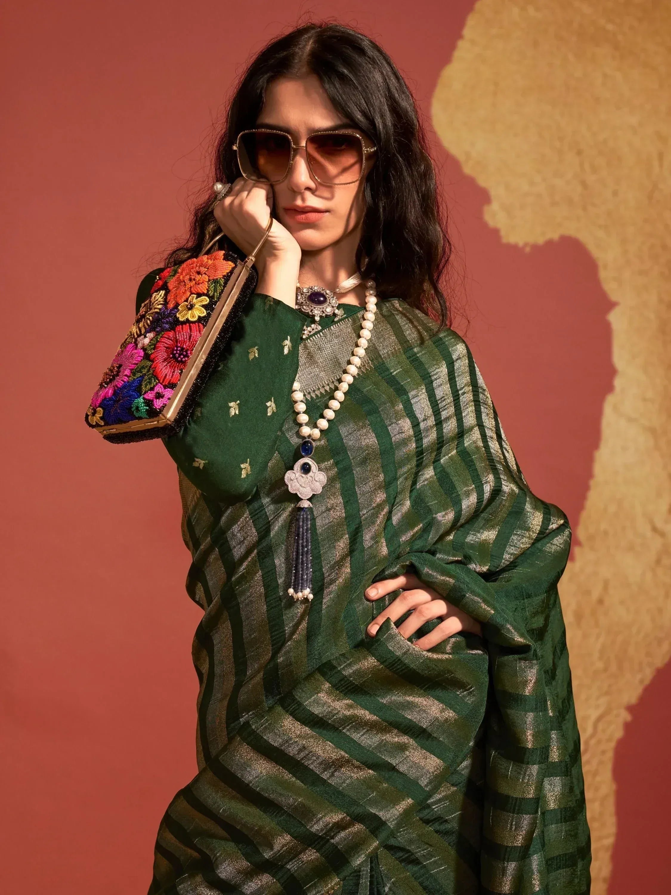 Woman in green striped saree with floral clutch, ethnic jewelry, and sunglasses, styled for Indian fashion.