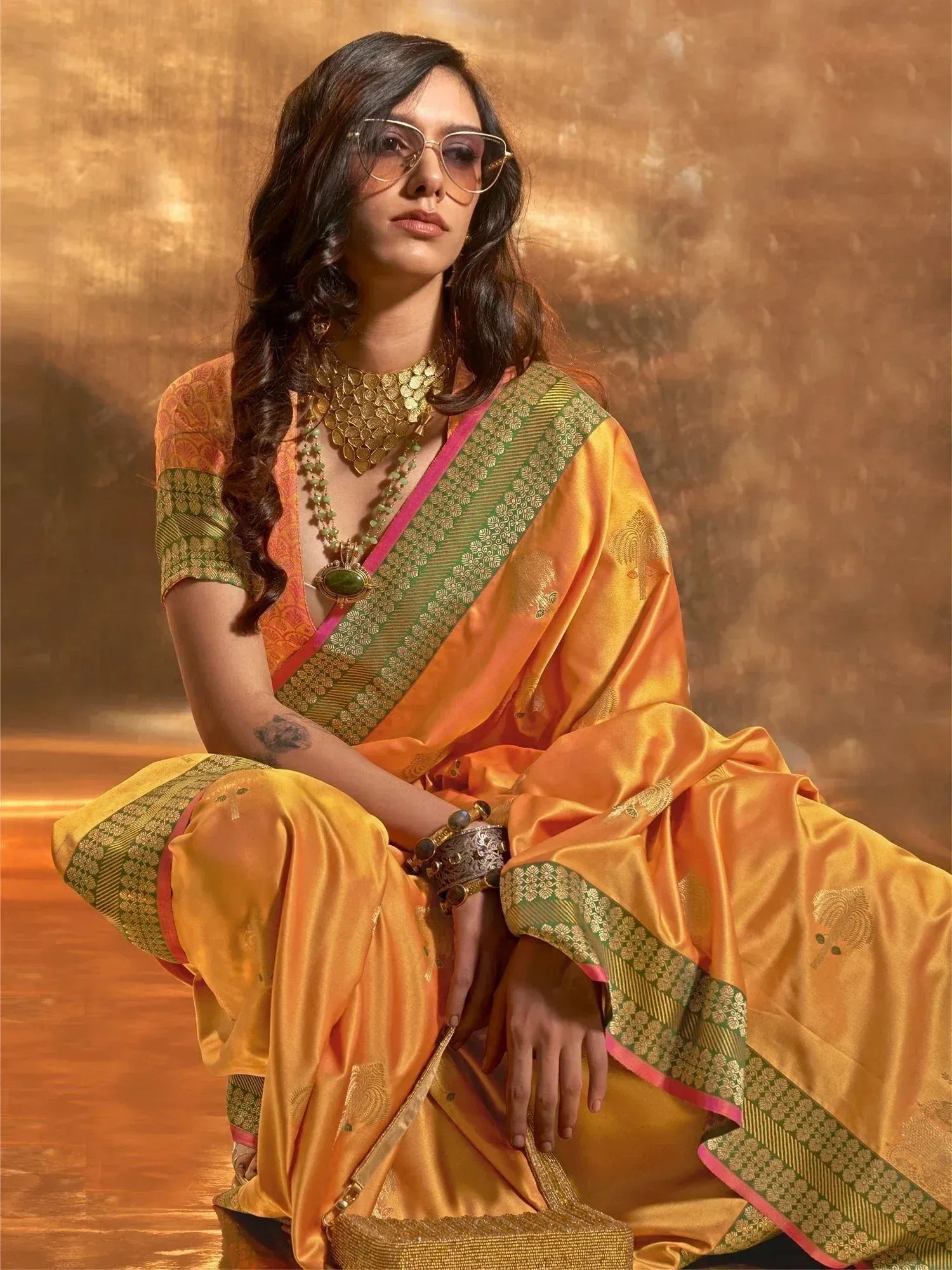 Woman in vibrant orange and green silk saree with gold embroidery, styled with statement jewelry and sunglasses