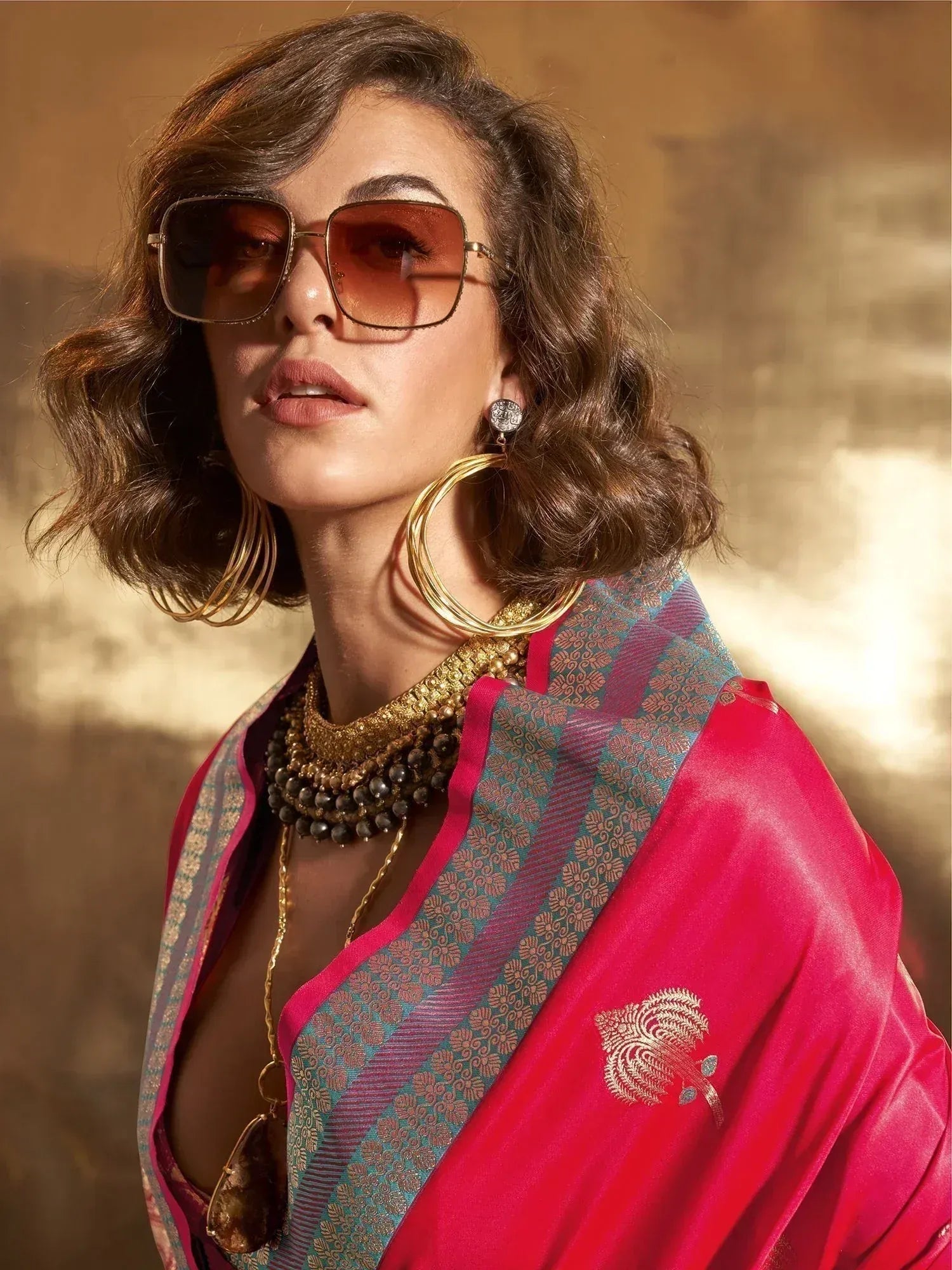 Stylish woman in vibrant pink ethnic saree with gold embroidery, statement jewelry, and sunglasses
