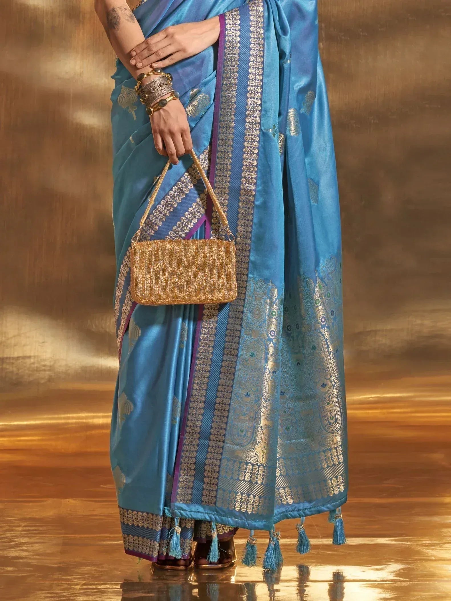 Blue silk saree with gold paisley border, tassels, and matching clutch for Indian ethnic wear