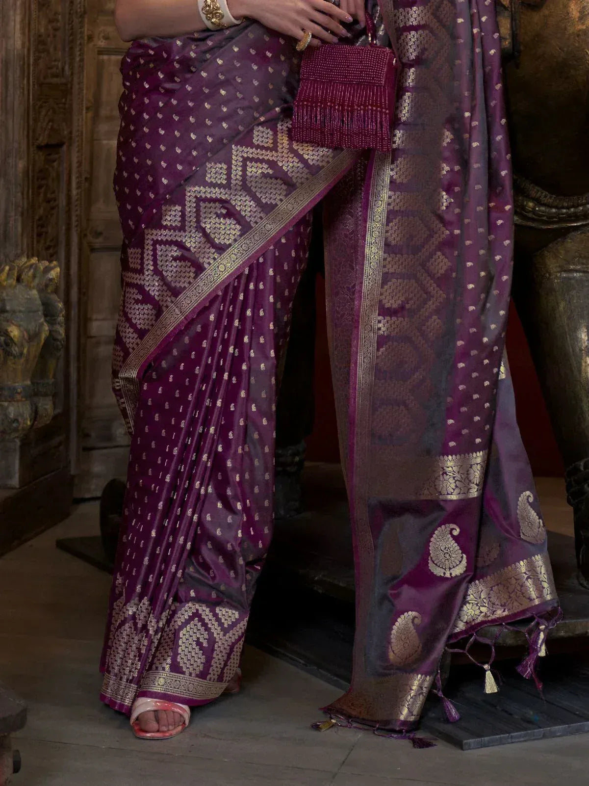 Purple silk saree with gold zari work, paisley motifs, and temple border, paired with matching clutch for ethnic wear.