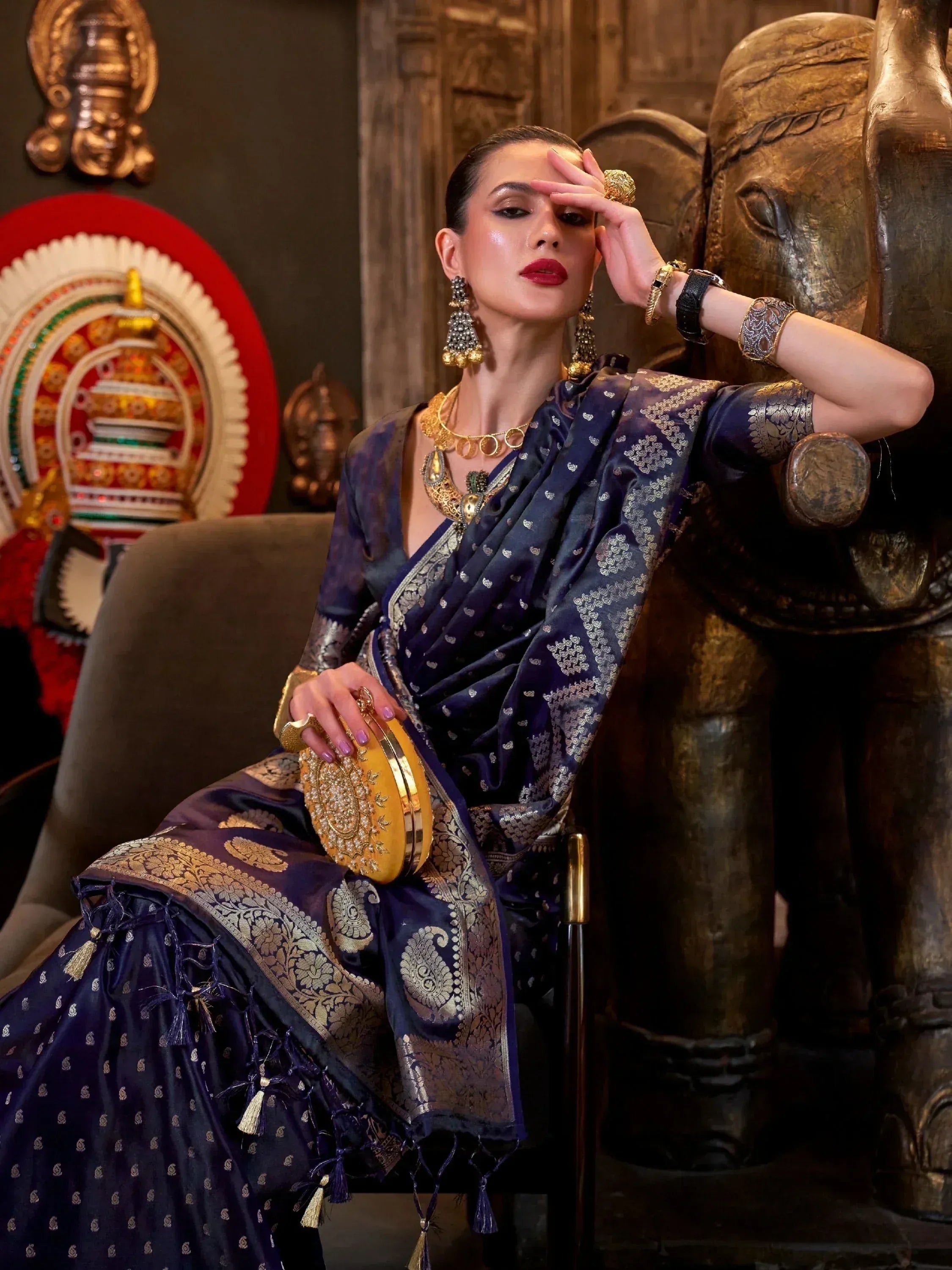 Elegant woman in navy blue designer saree with gold embroidery, traditional jewelry, and clutch, seated beside elephant statue.