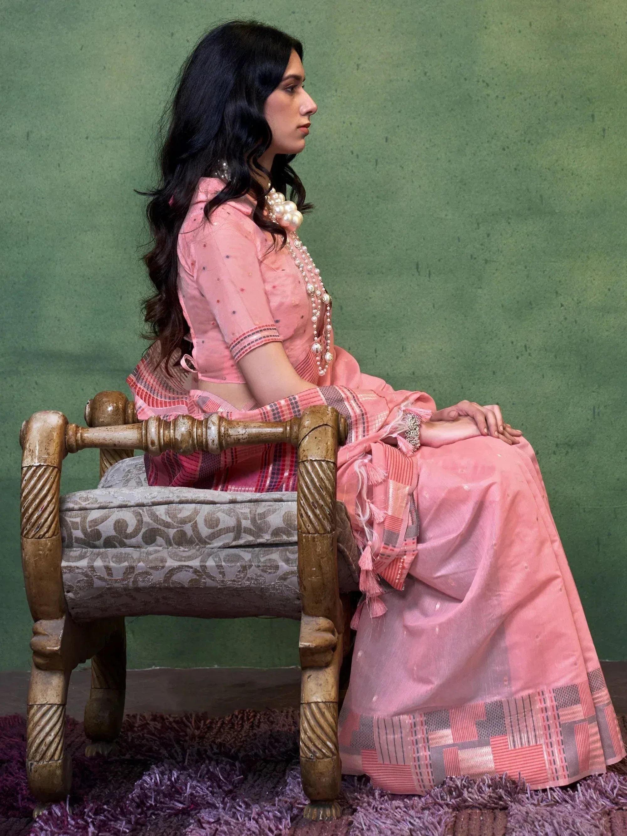 Woman in pink ethnic lehenga choli with pearl jewelry, seated on carved chair against green backdrop