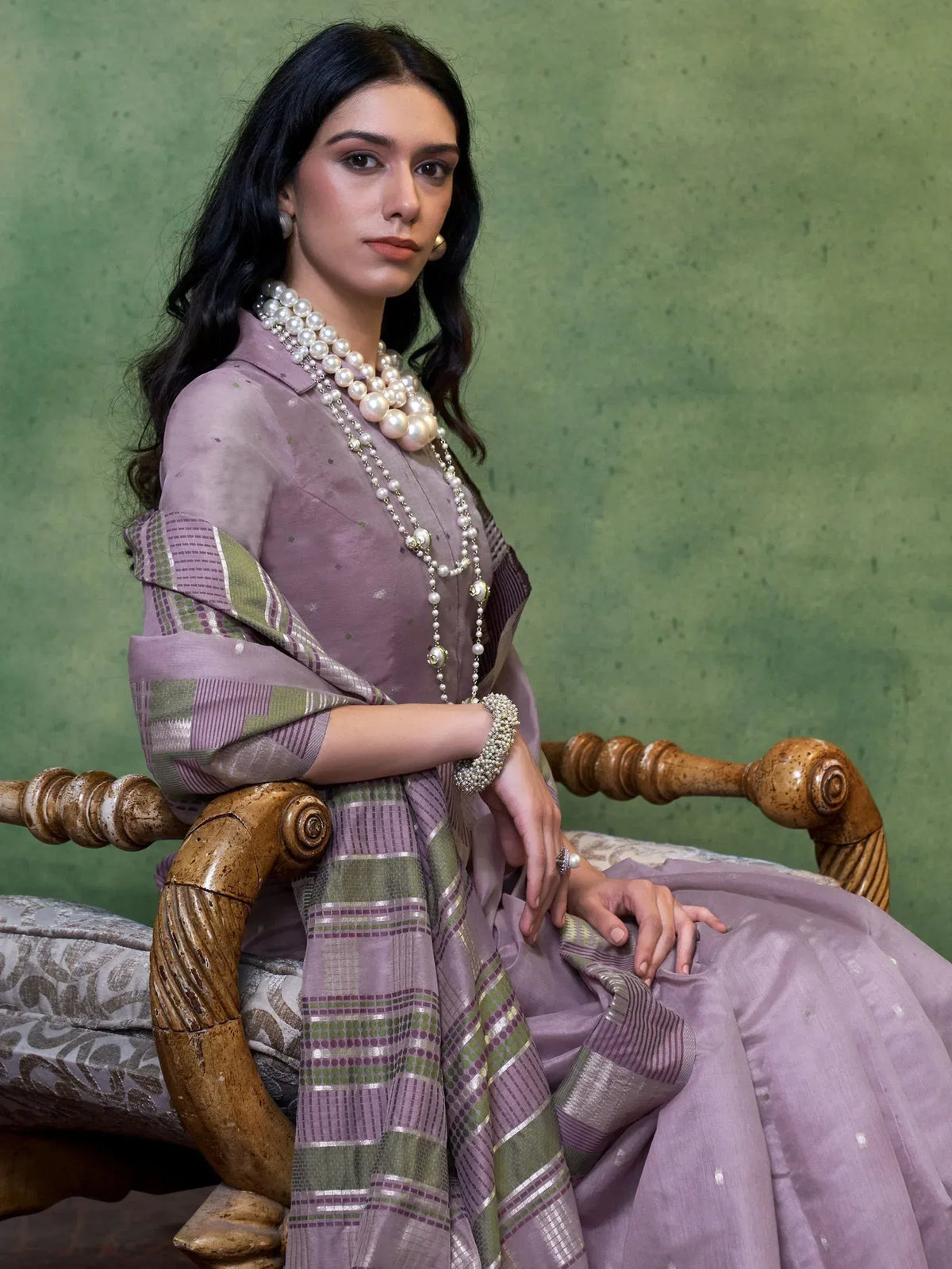 Woman in elegant lavender ethnic gown with pearl jewelry, seated on wooden chair against green backdrop