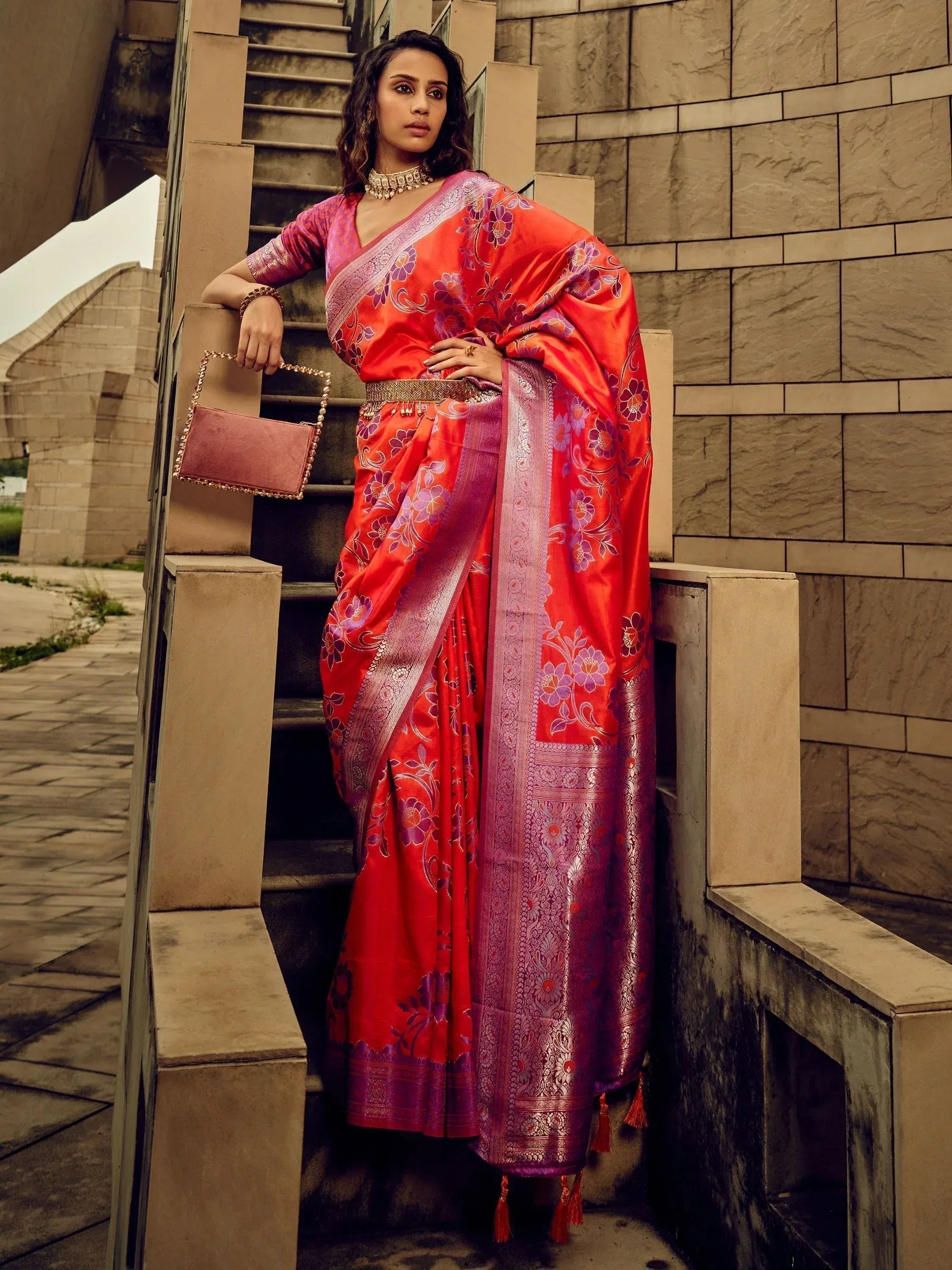 Elegant woman in vibrant red floral saree with gold accents, styled with a matching clutch and jewelry.