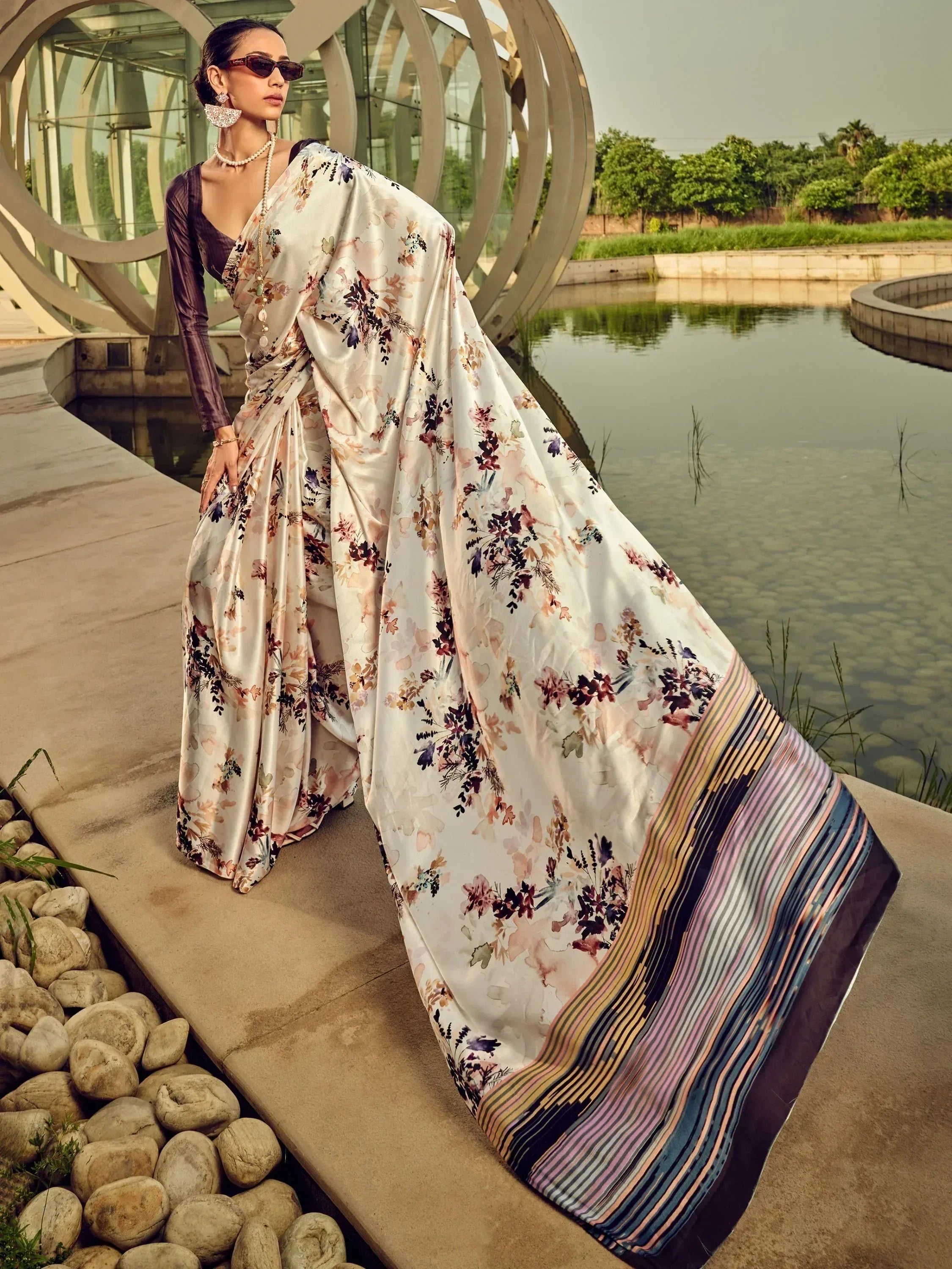 Woman in floral print saree with striped border, styled with sunglasses and pearl jewelry, near water feature