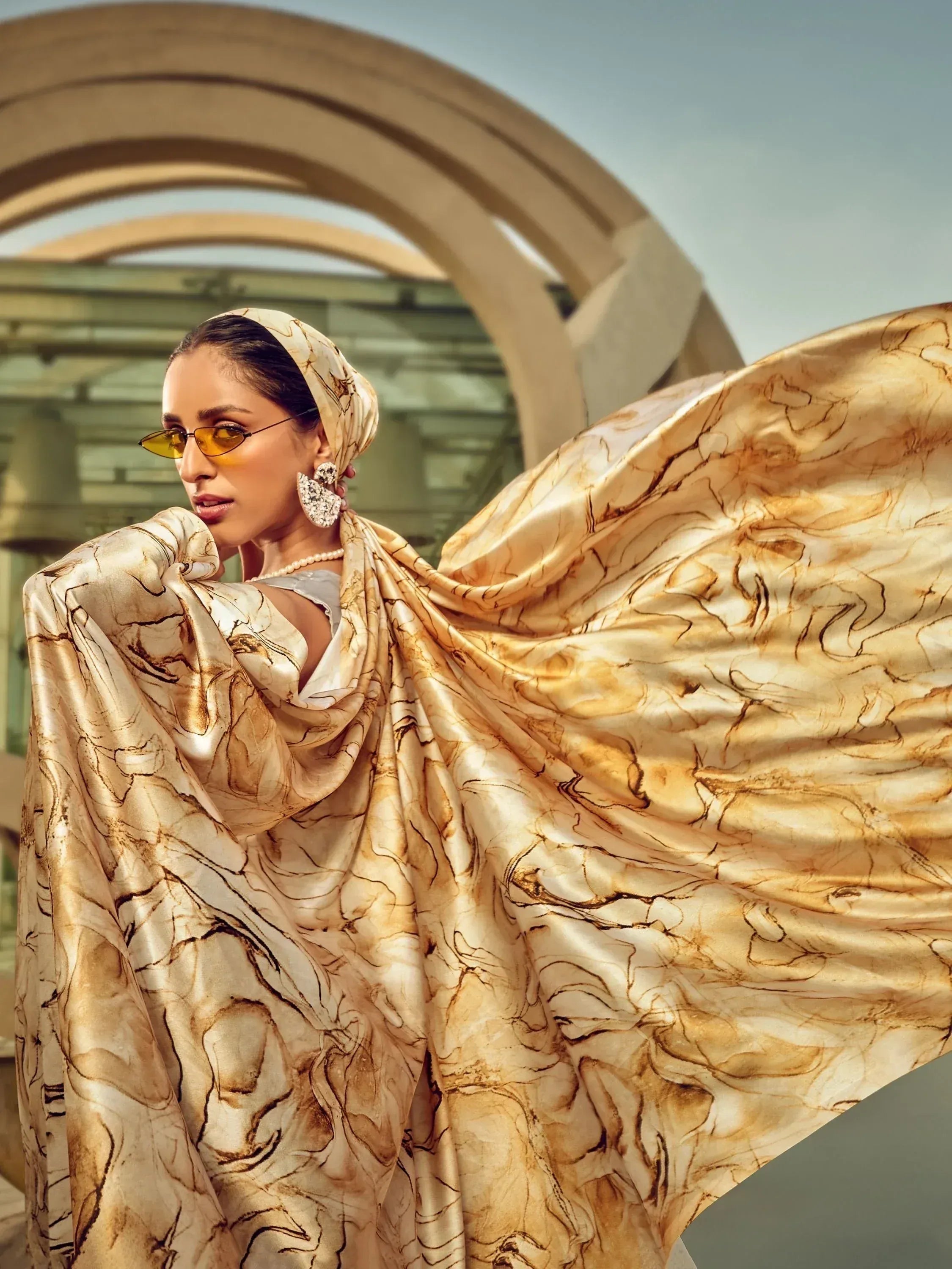 Woman in flowing gold marble print ethnic gown with headscarf, sunglasses, and statement earrings