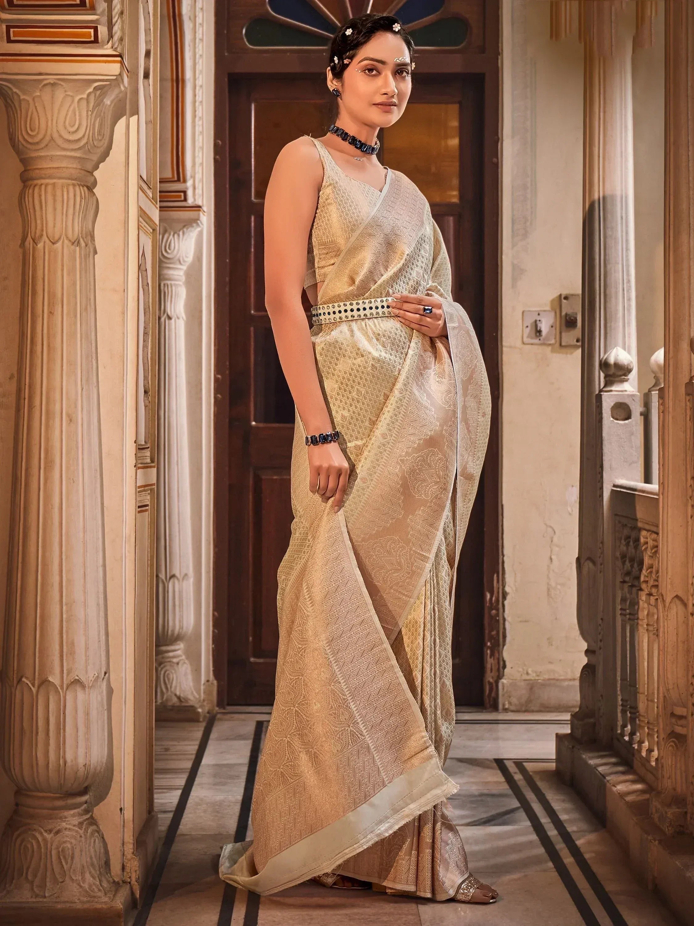 Elegant woman in gold embroidered saree with blue jewelry, standing in ornate Indian hallway