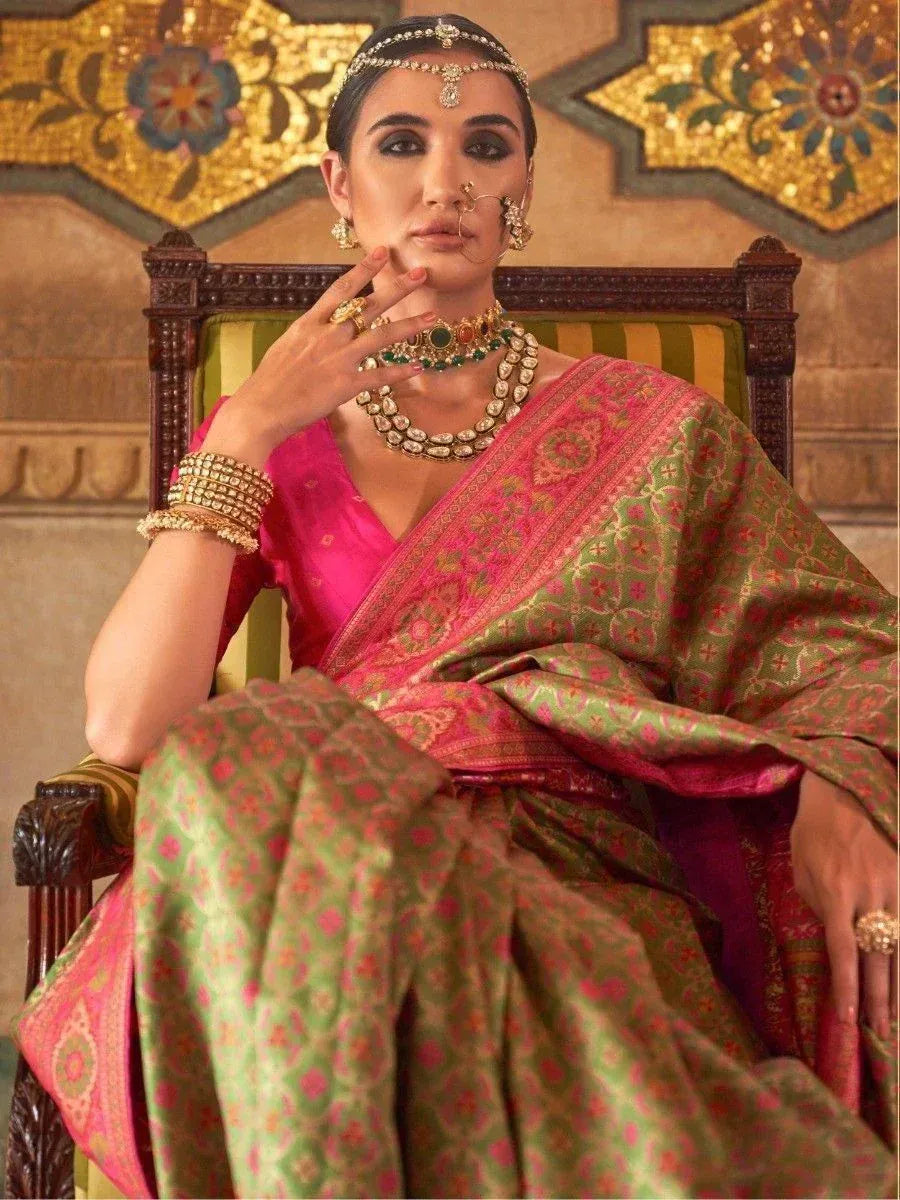 Woman in pink and green designer saree with traditional jewelry, seated on ornate chair