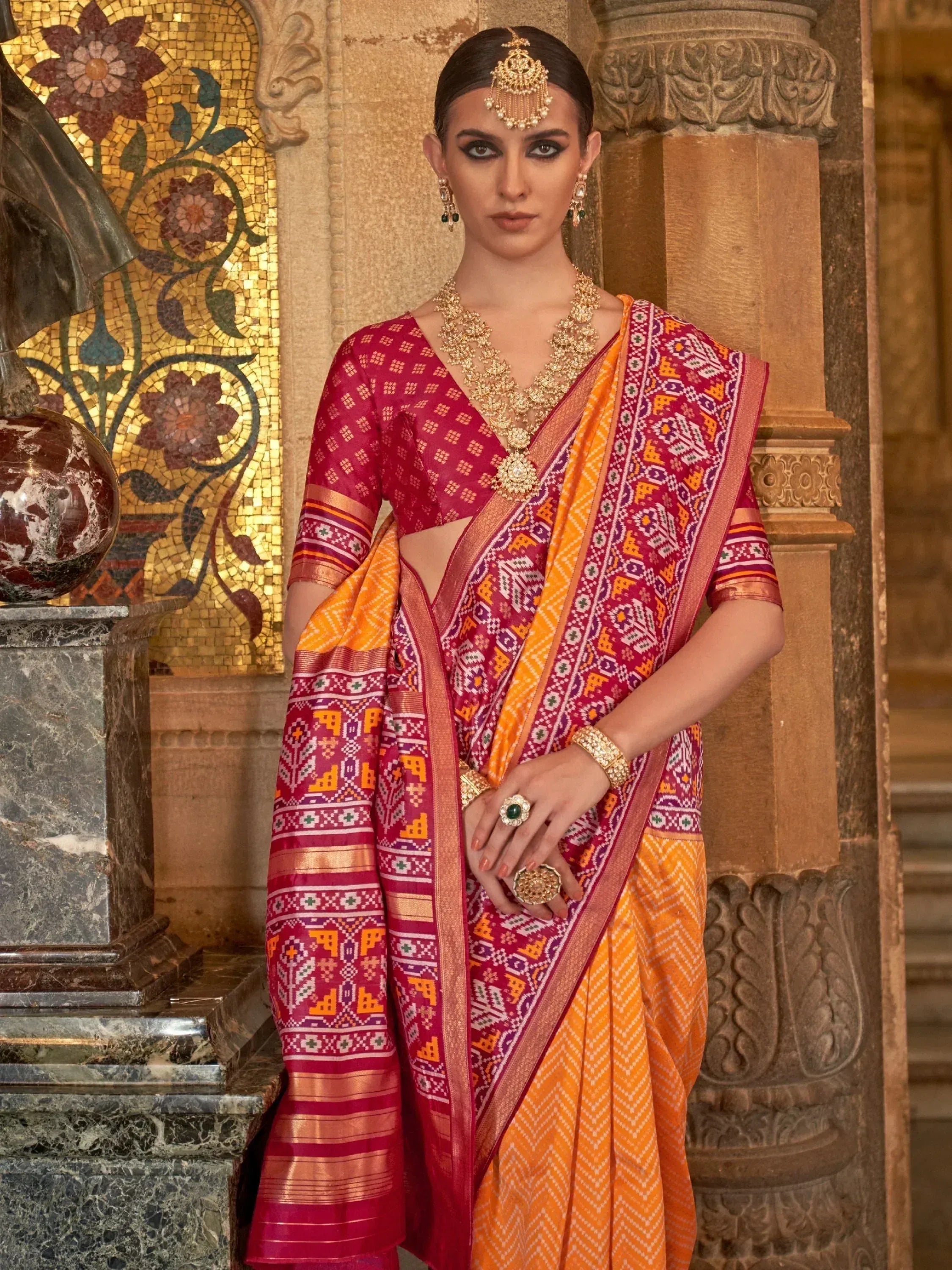 Vibrant red and orange ethnic saree with geometric patterns, paired with gold jewelry, perfect for Indian weddings or festivals.