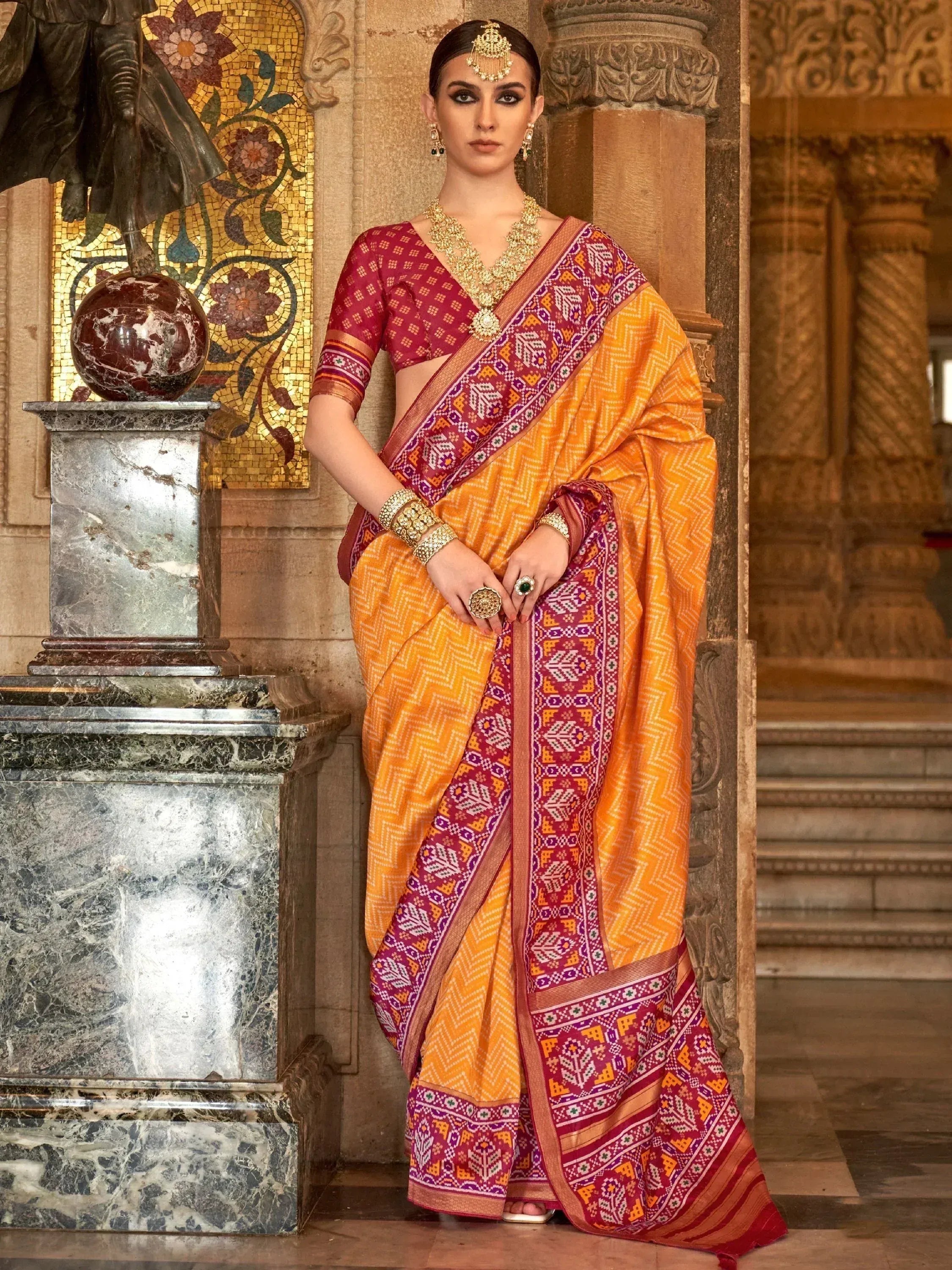 Woman in vibrant orange and maroon ethnic saree with intricate patterns, traditional jewelry, palace backdrop