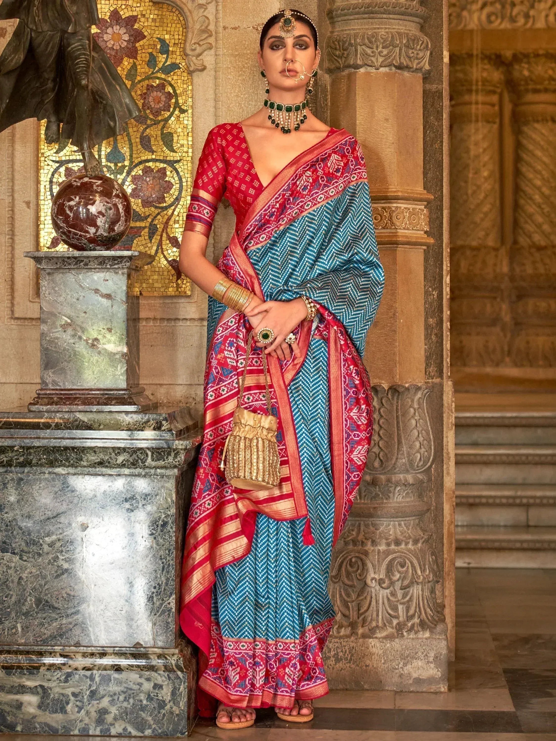 Woman in red and blue printed saree with gold jewelry, standing near ornate pillar and mosaic wall
