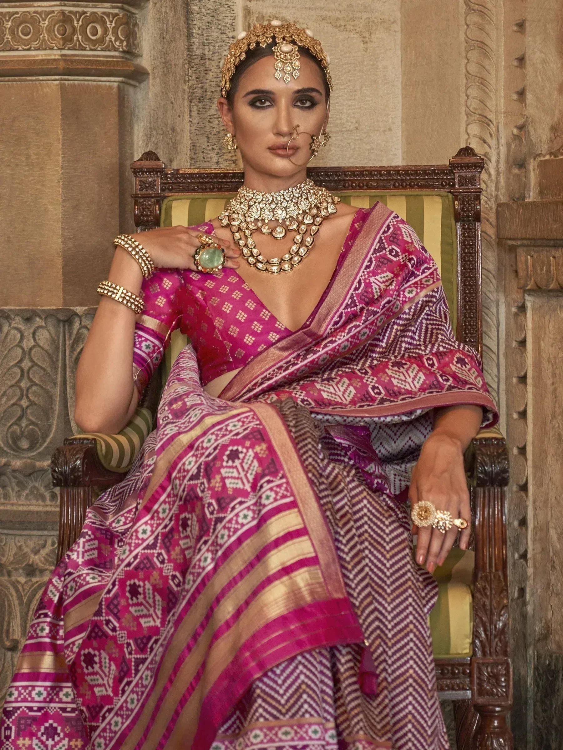 Woman in vibrant pink ethnic saree with gold jewelry, seated on ornate chair, traditional Indian attire