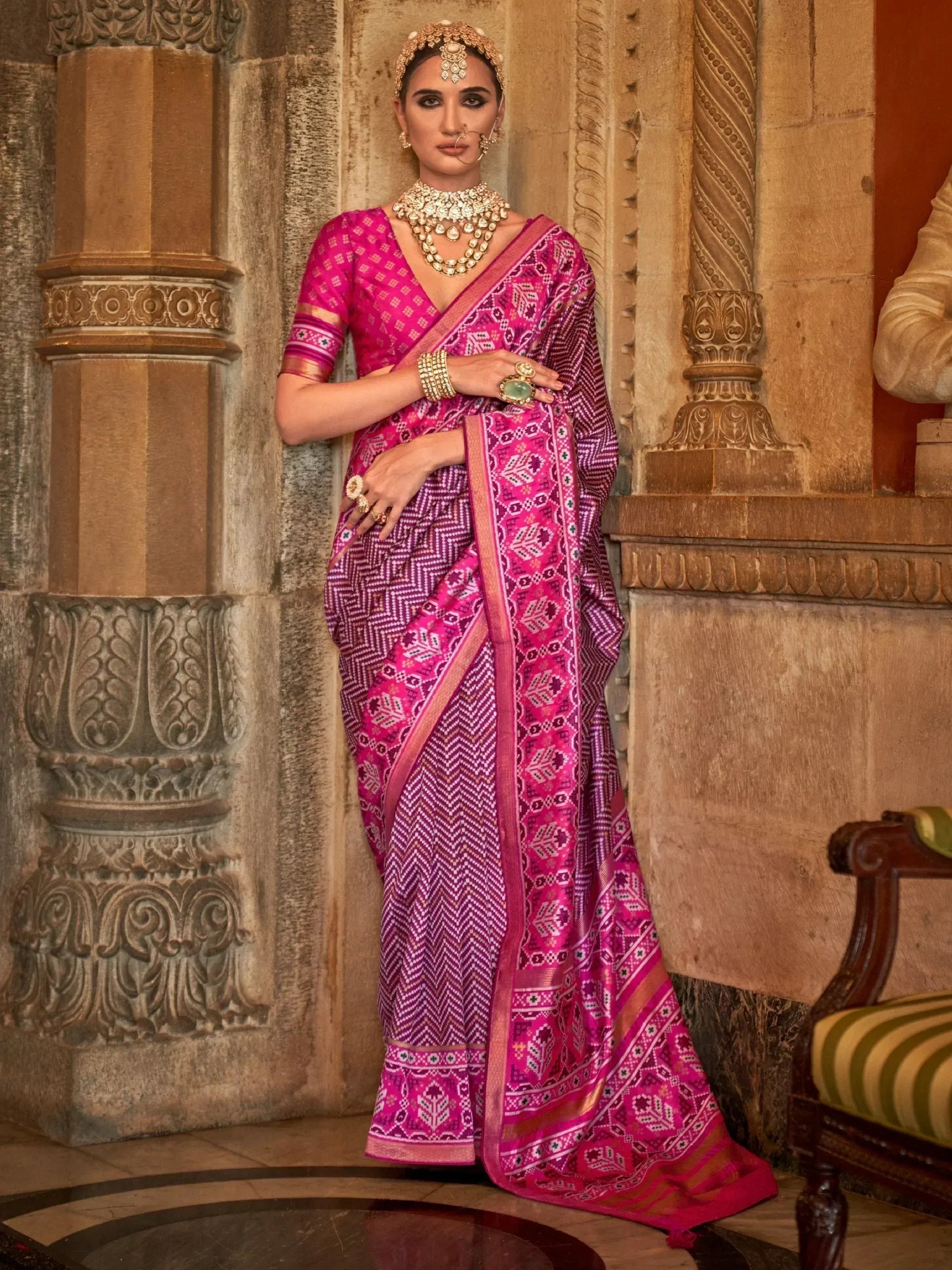 Woman in vibrant pink ethnic saree with geometric patterns, traditional jewelry, and ornate temple backdrop