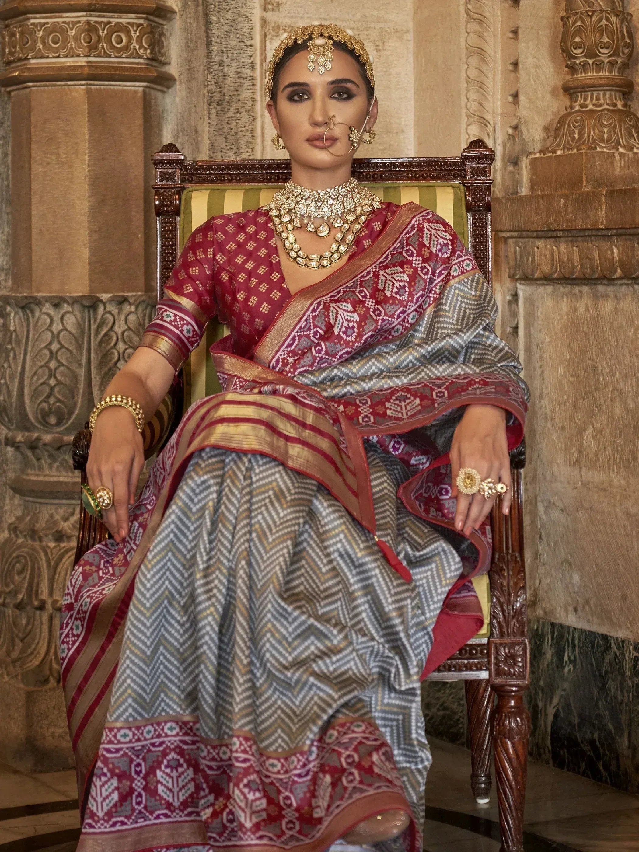 Woman in traditional Indian saree with intricate patterns, gold jewelry, seated on ornate chair