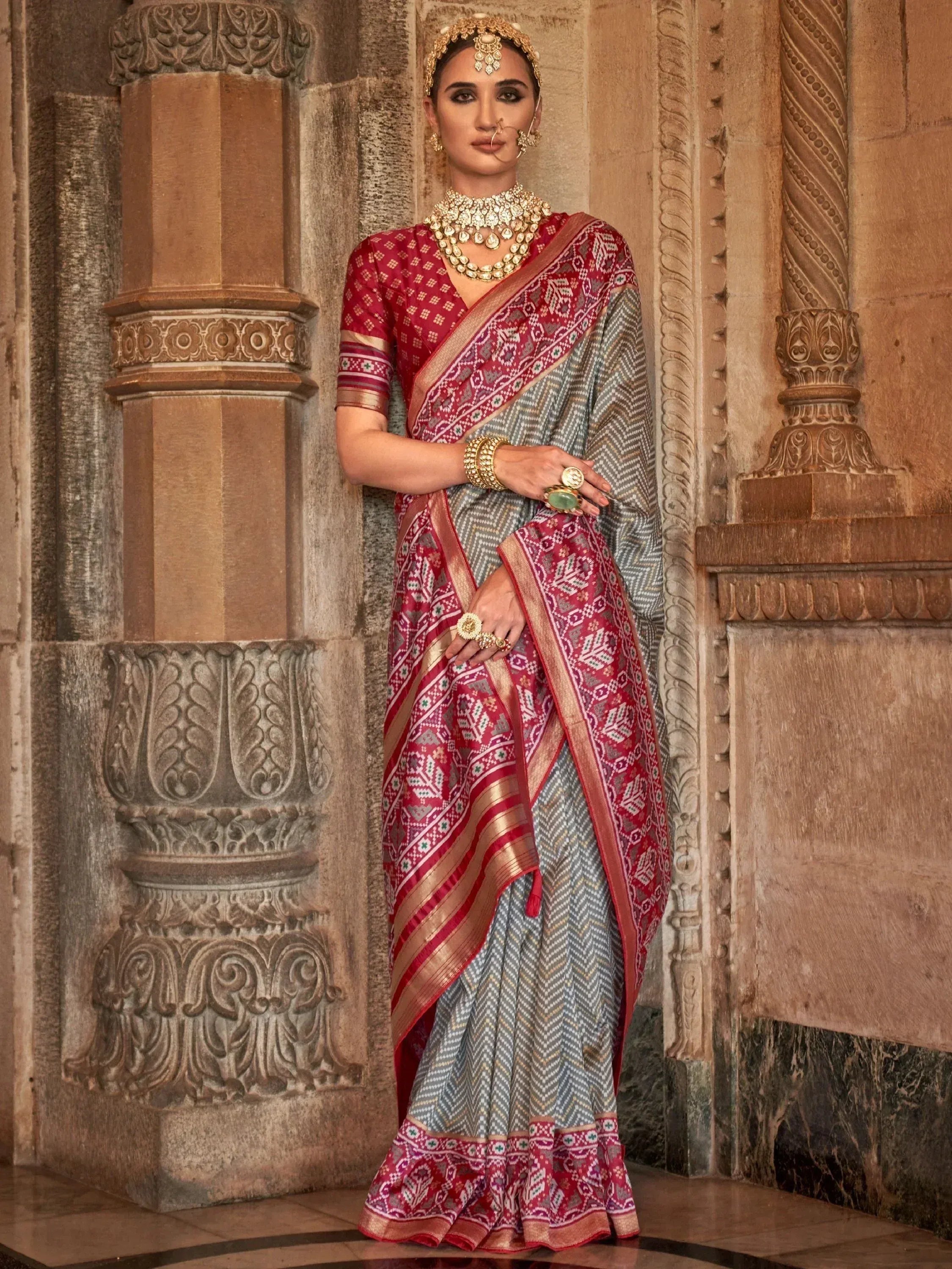 Woman in red and grey printed saree with gold jewelry, standing by carved stone pillars