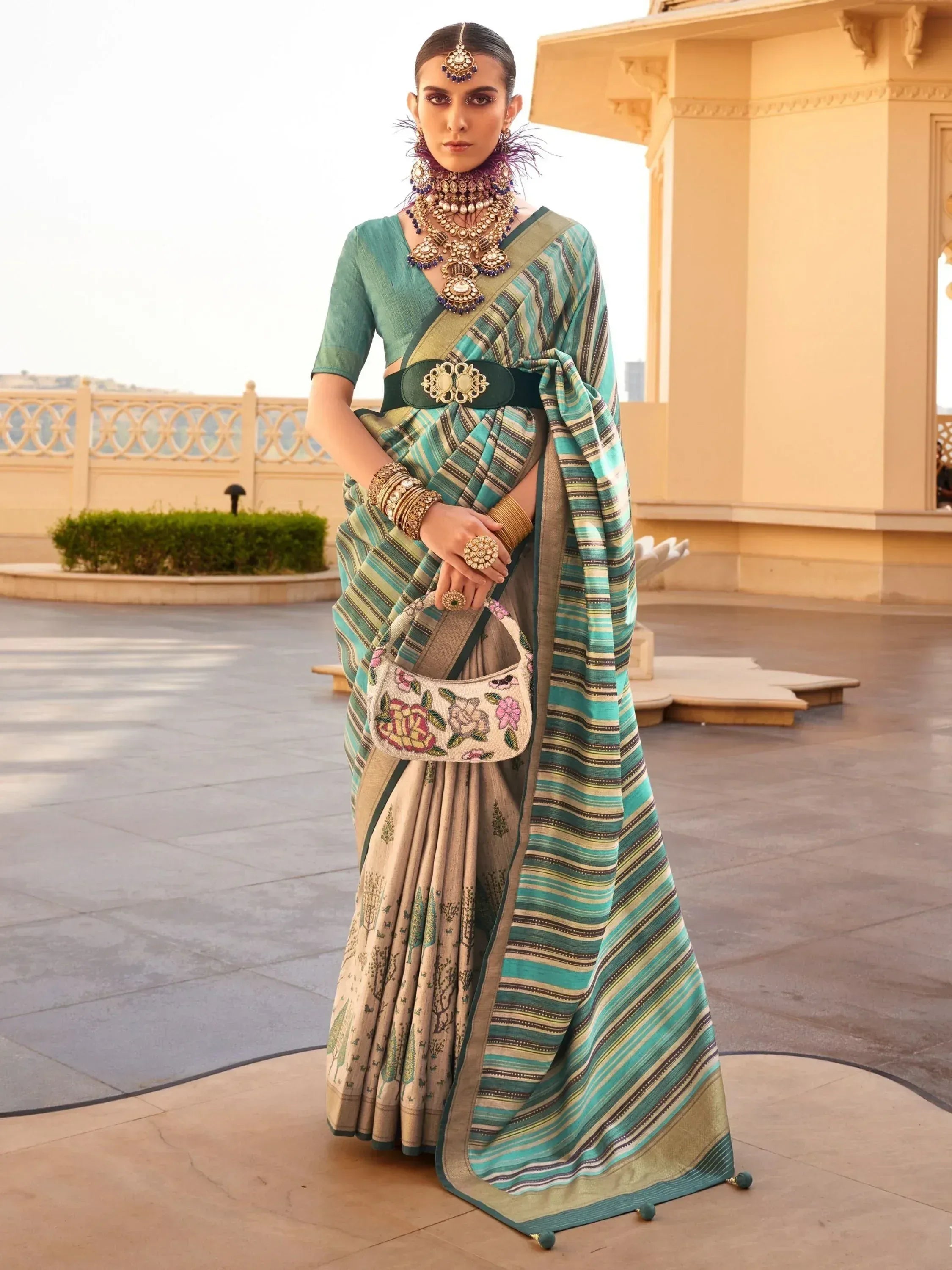 Woman in designer saree with striped pallu, ethnic jewelry, and floral handbag, perfect for Indian occasions.