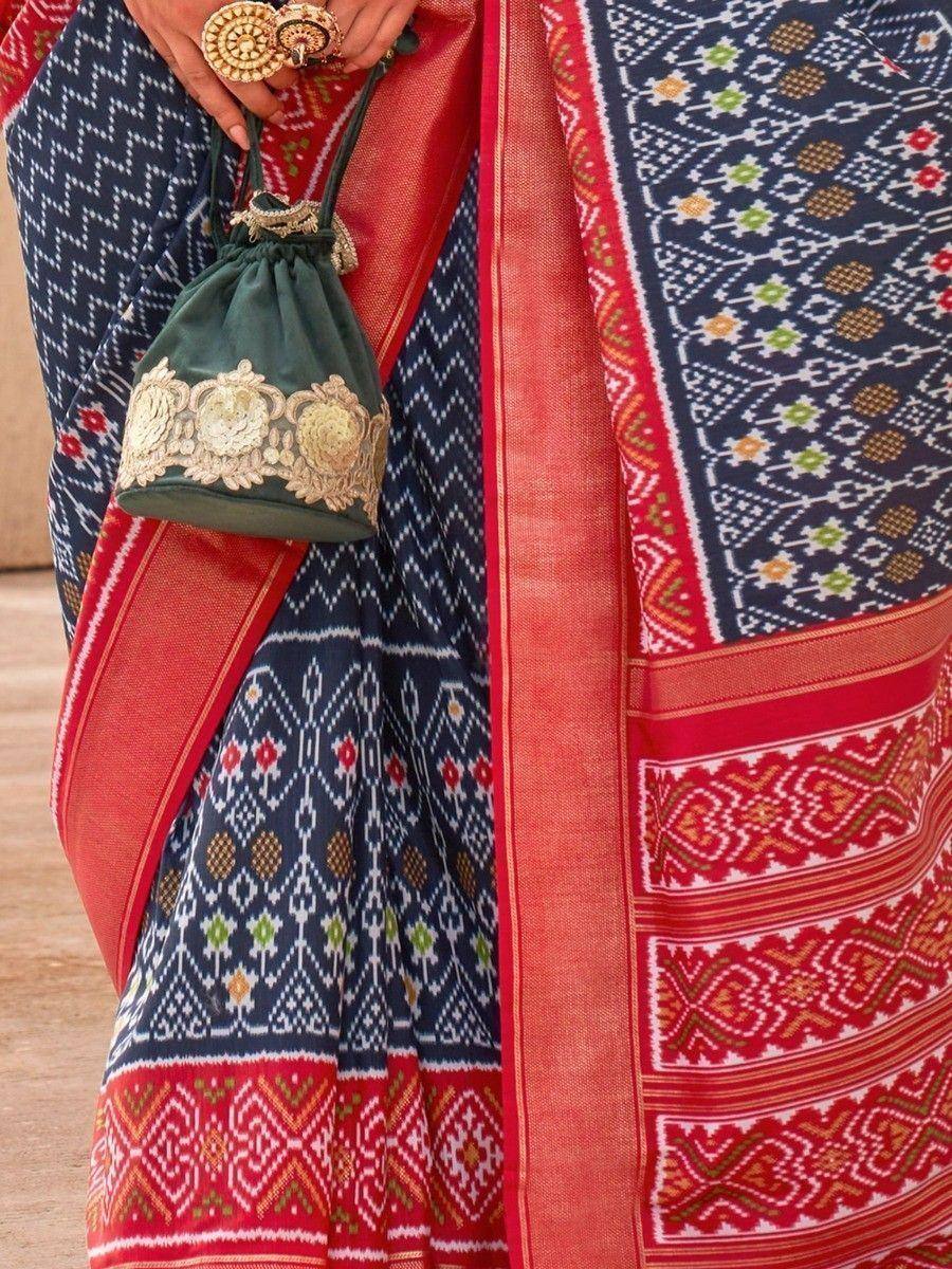 Blue and red patterned ethnic saree with gold embroidery, paired with a green potli bag and traditional jewelry.