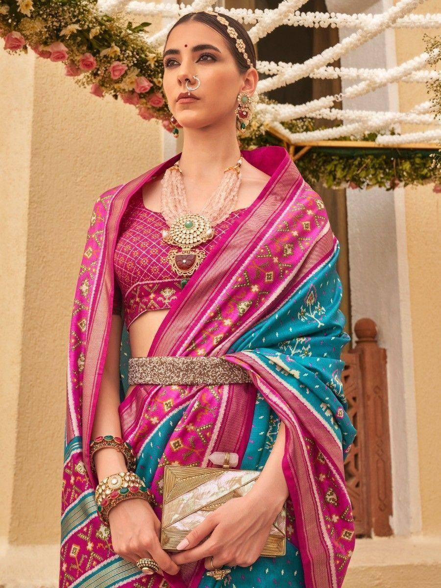 Pink and teal designer saree with gold embroidery, traditional jewelry, and floral backdrop