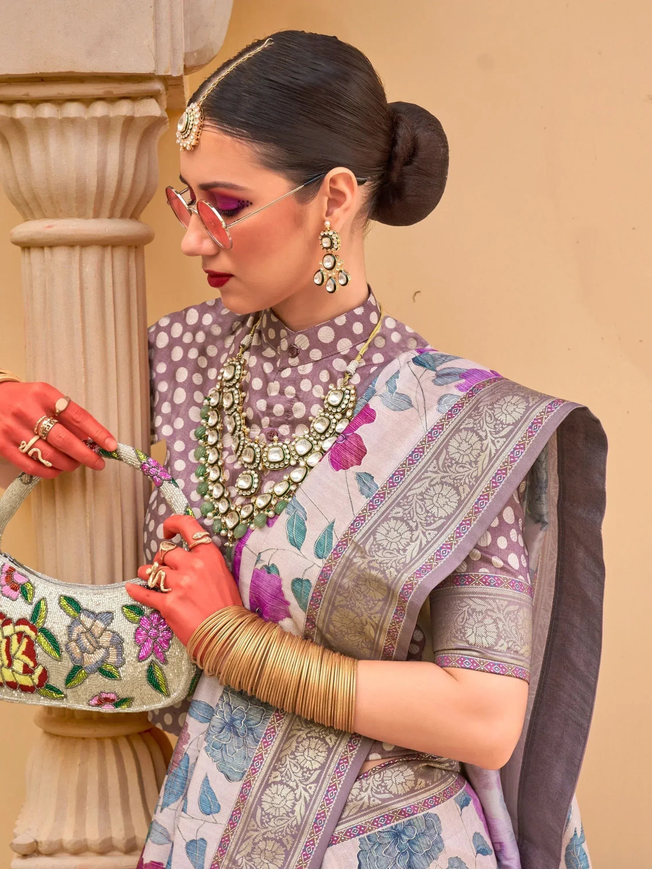 Woman in floral saree with polka dot blouse, traditional jewelry, and beaded handbag, styled for ethnic occasions.