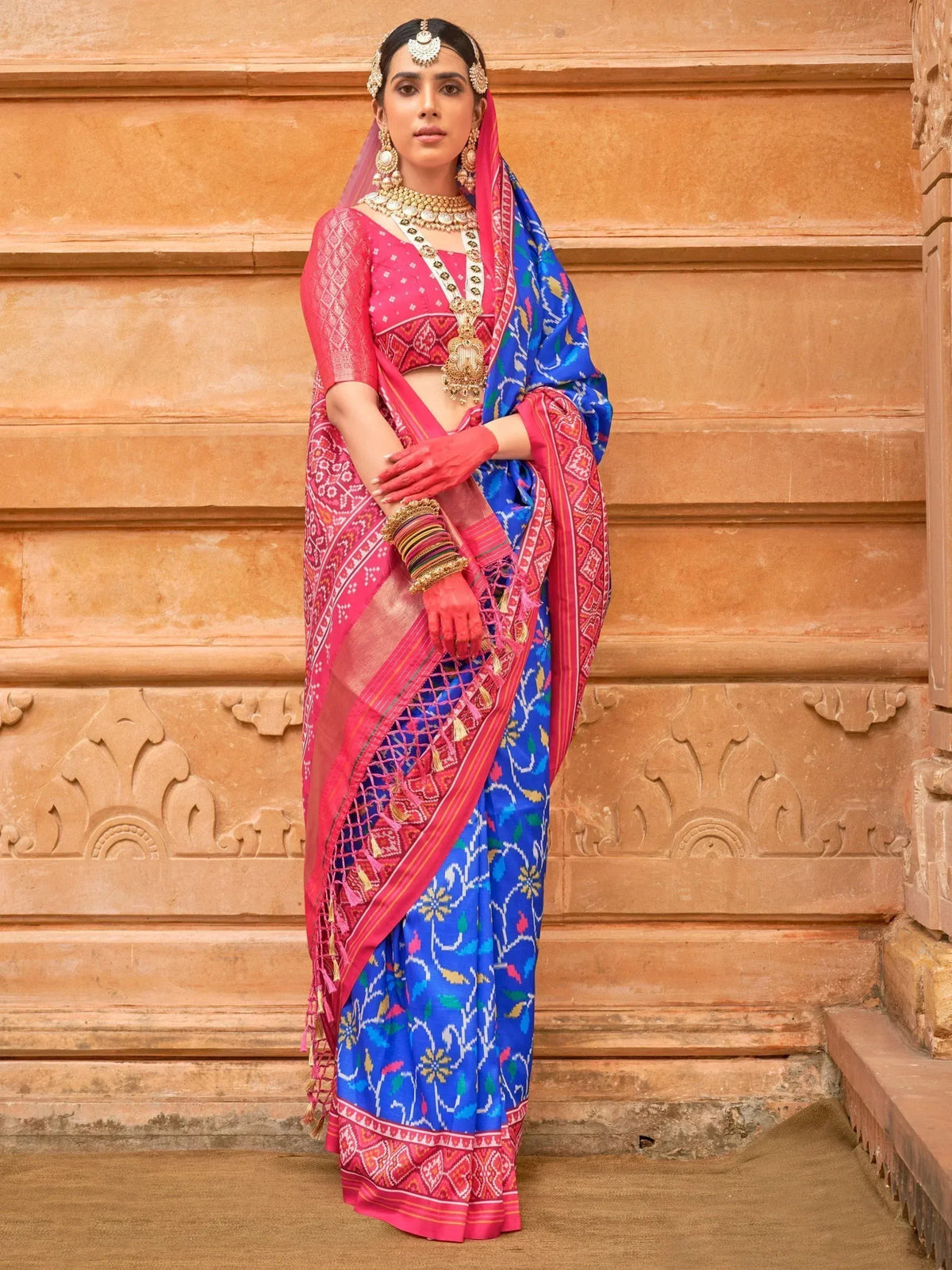 Woman in vibrant pink and blue Indian saree with traditional jewelry, standing against ornate sandstone wall