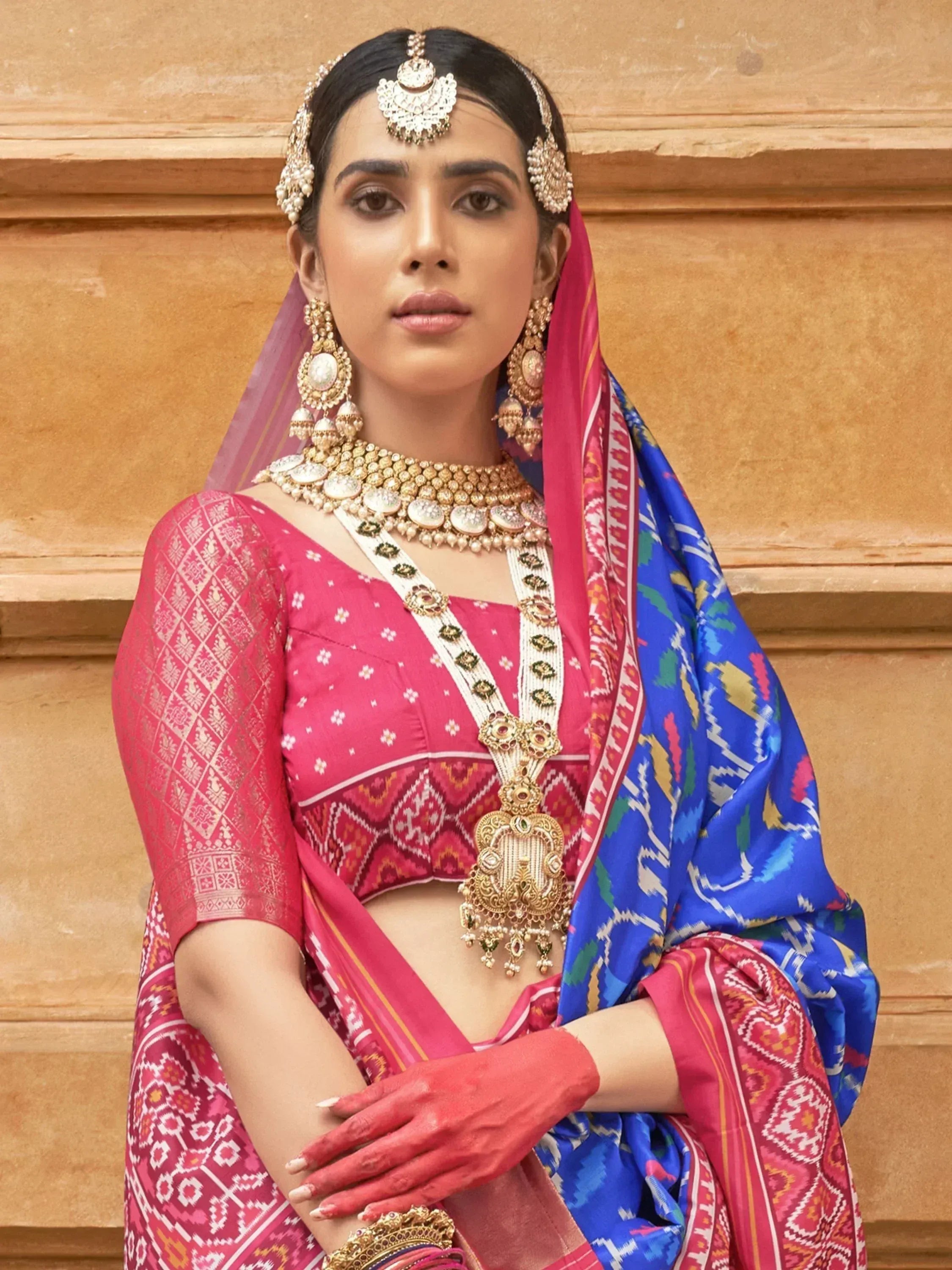 Woman in vibrant pink and blue ethnic saree with intricate patterns, traditional jewelry, and henna hands.