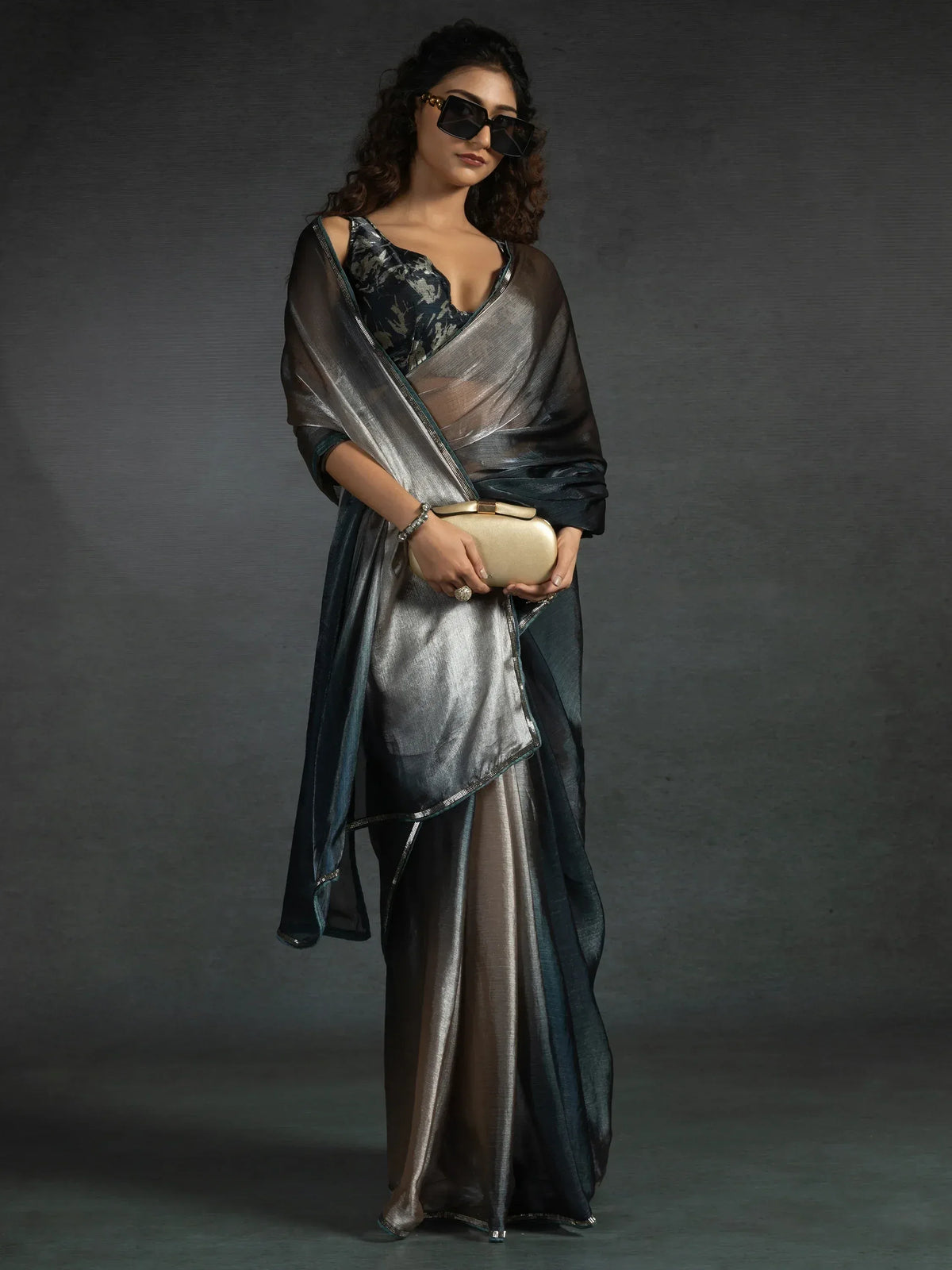Stylish woman in metallic grey and teal saree with floral blouse, holding gold clutch, wearing sunglasses