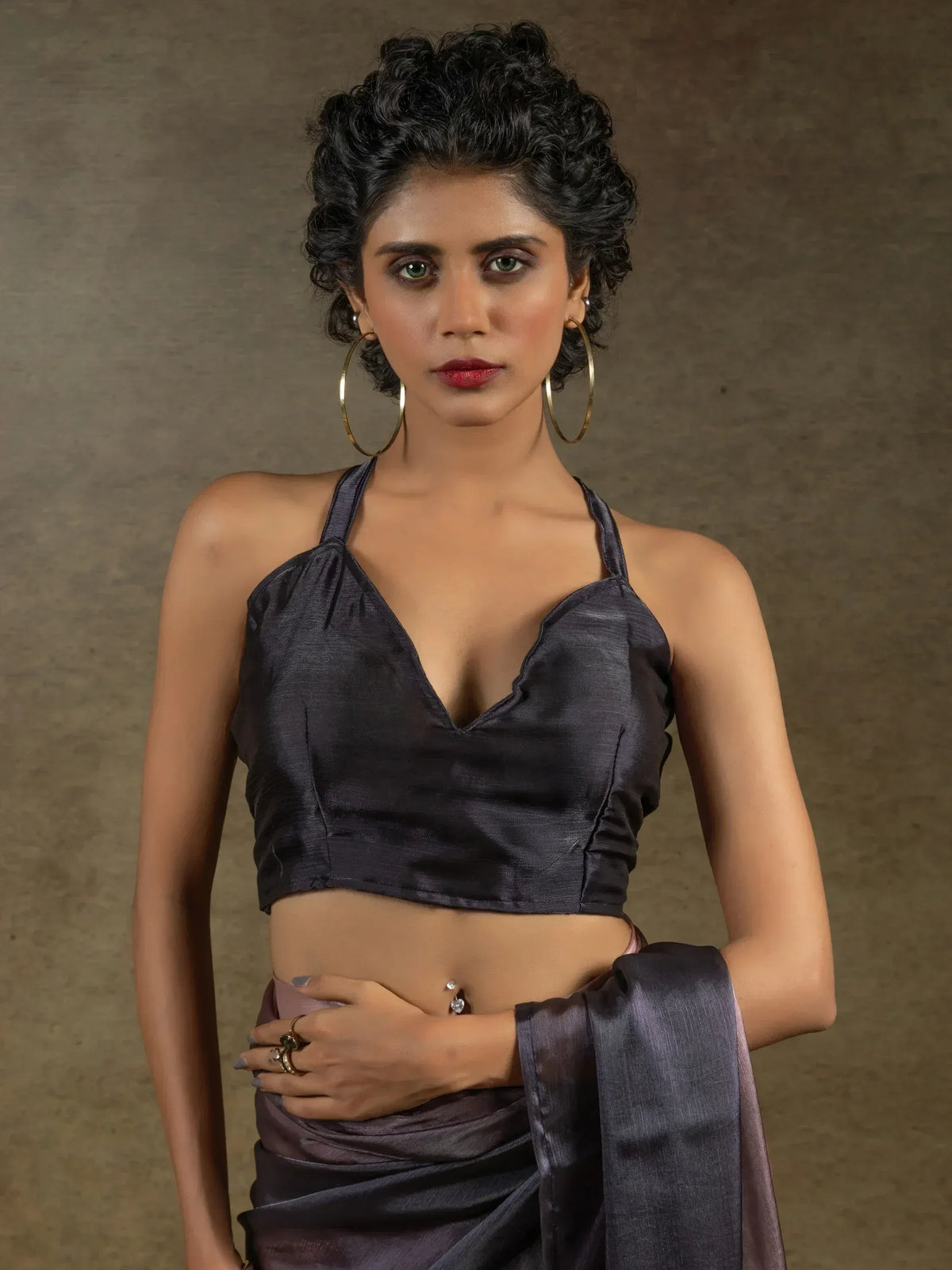 Woman in dark grey satin saree with halter neck blouse, gold hoops, and bold makeup
