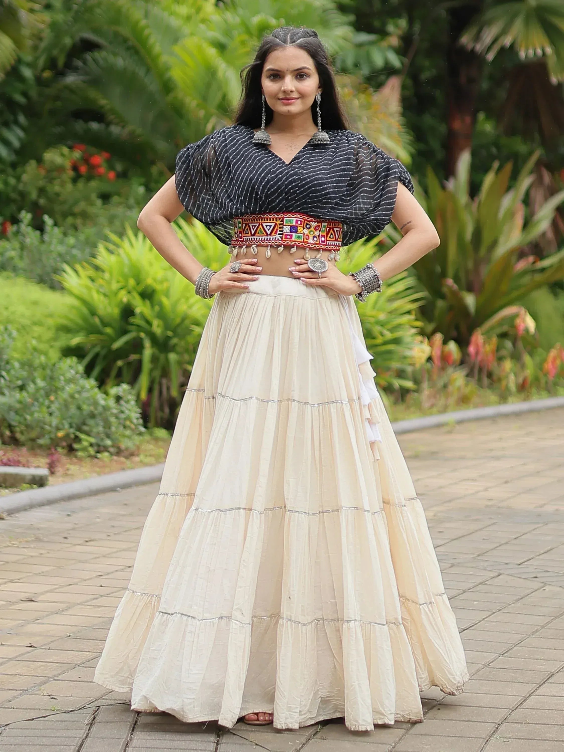 Woman in ethnic Indian outfit: black striped top, embroidered belt, cream tiered skirt, garden setting