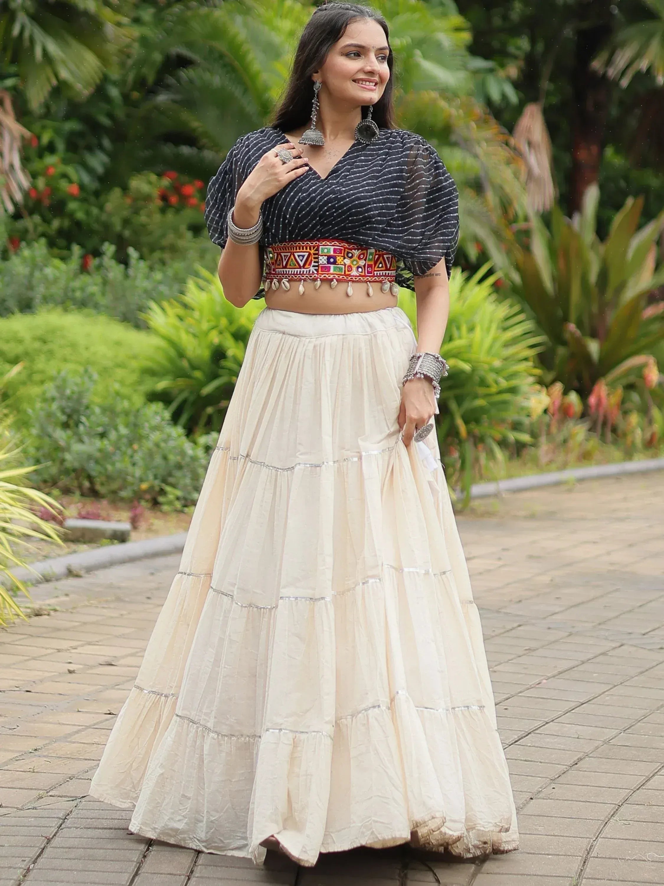 Woman in ethnic Indian outfit with striped crop top, embroidered belt, and tiered cream skirt, styled with traditional jewelry