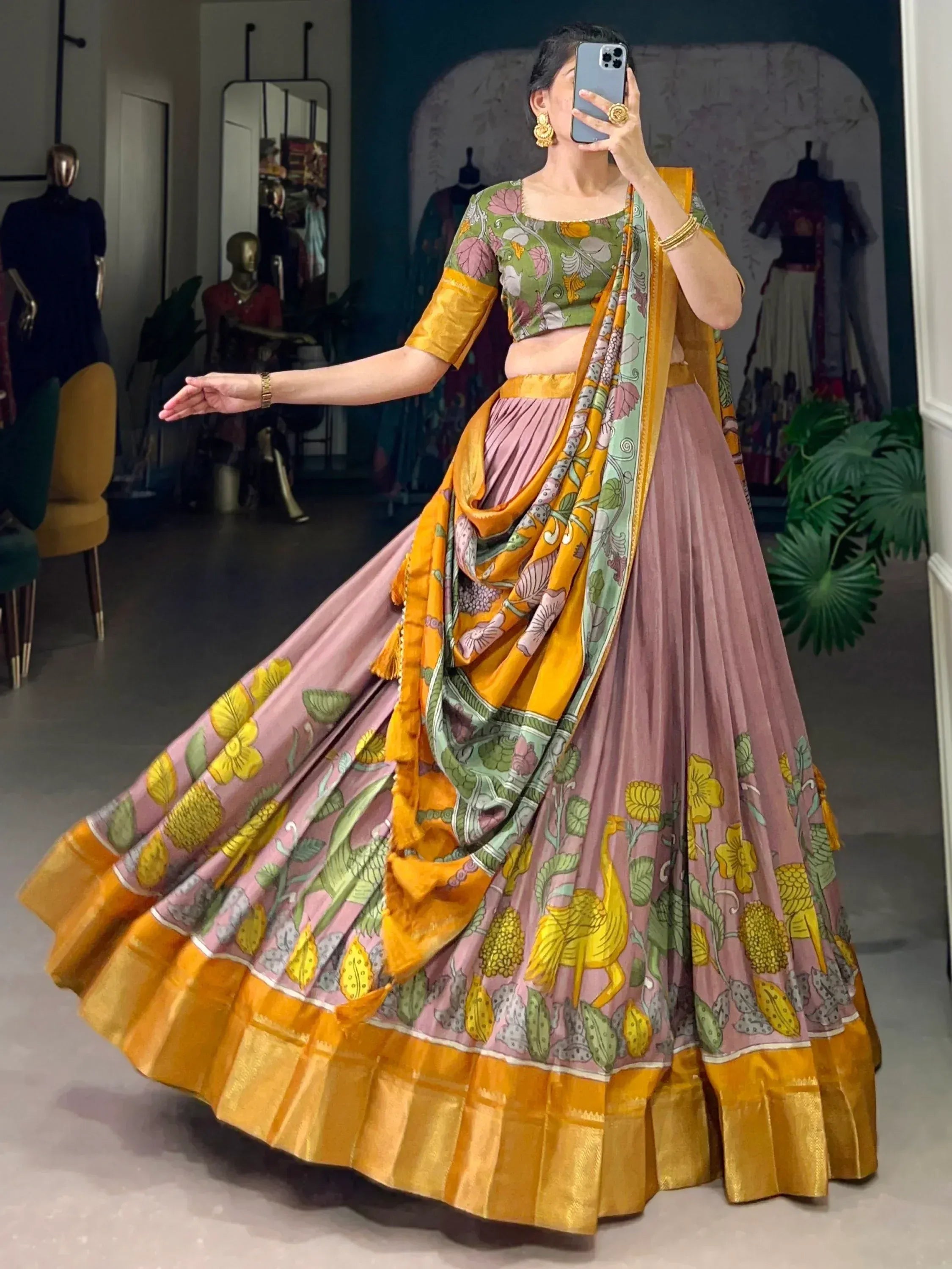Woman in floral print lehenga choli with pink skirt, green blouse, and yellow dupatta, taking mirror selfie in boutique