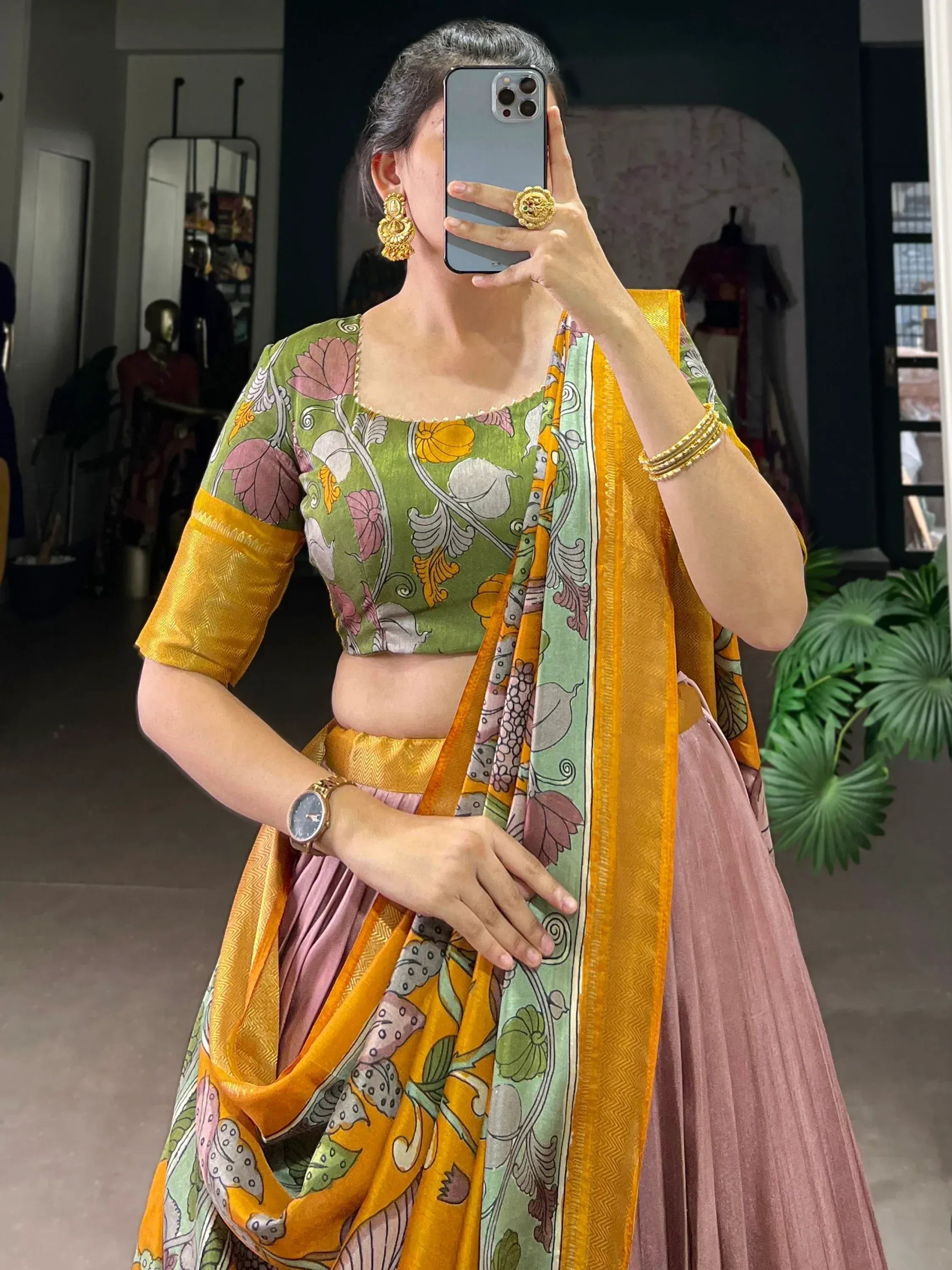 Woman in floral green and yellow lehenga choli with gold jewelry, taking mirror selfie