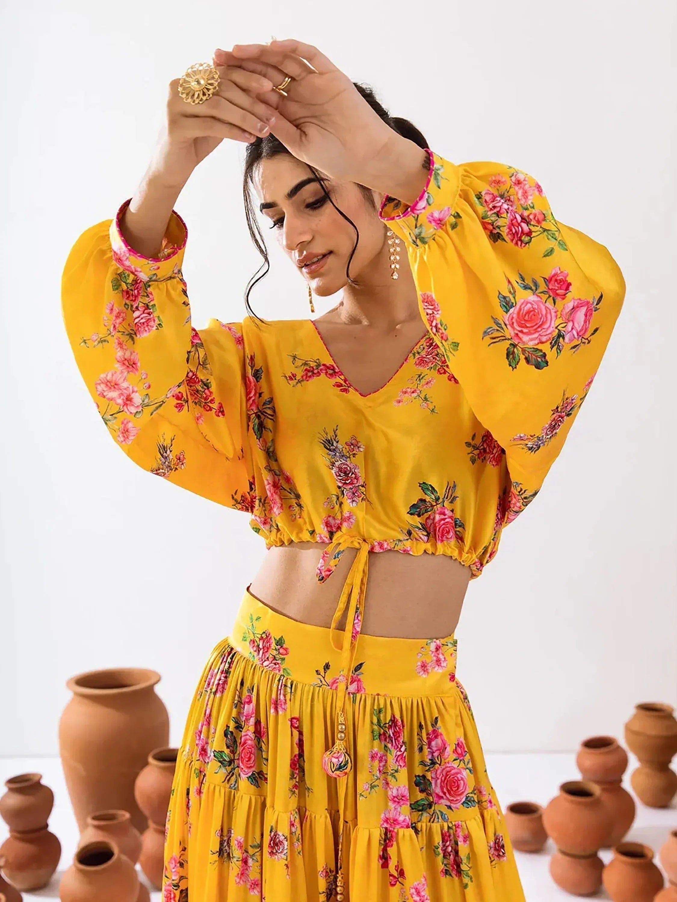 Yellow floral lehenga choli with puff sleeves and tie-up crop top, styled with gold jewelry and clay pots.