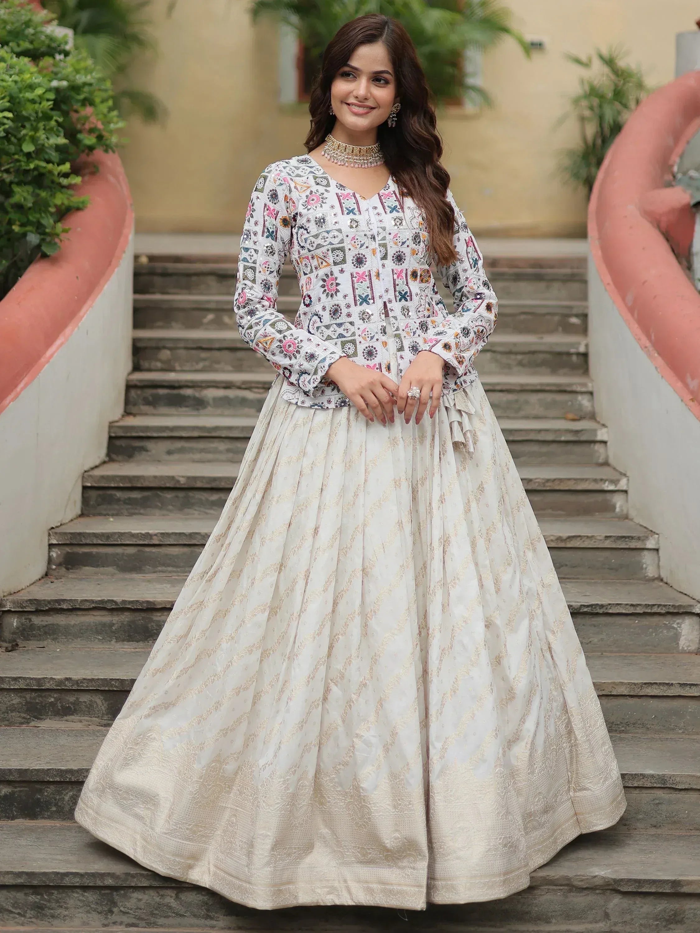 Woman in white embroidered Indian ethnic wear with floral top and gold-patterned skirt, standing on stairs
