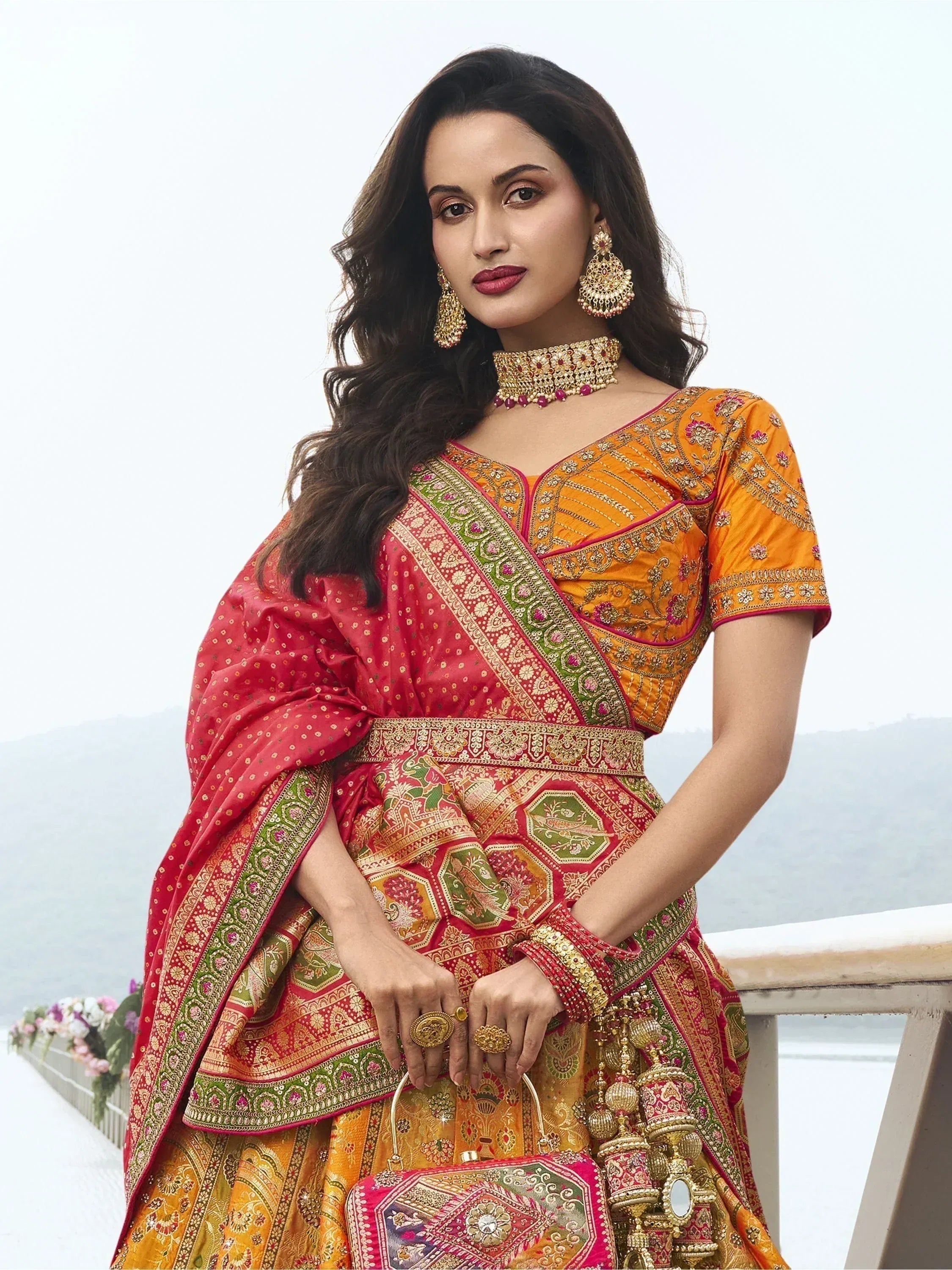 Woman in vibrant orange and red embroidered lehenga choli with gold jewelry, perfect for Indian ethnic occasions.