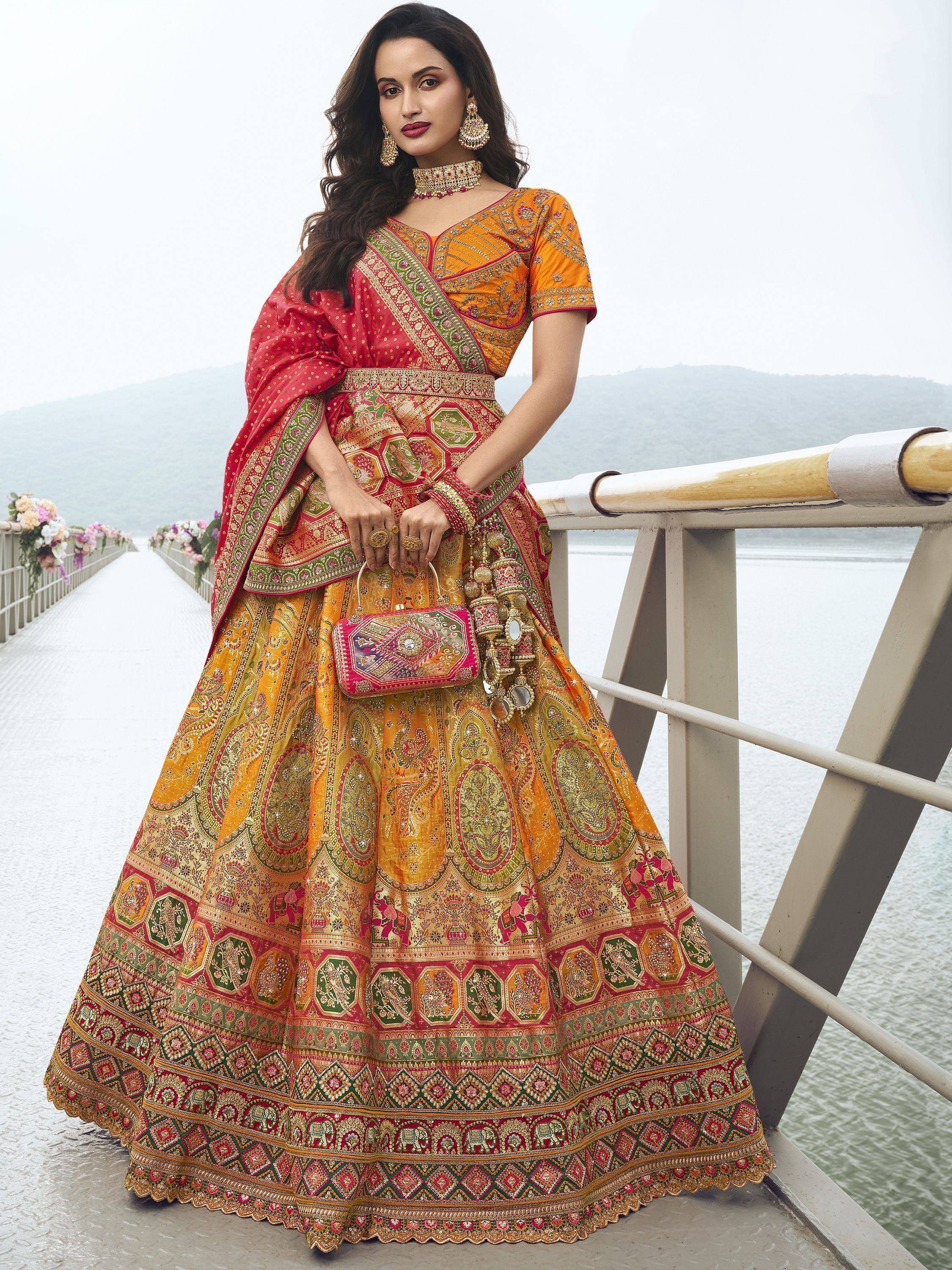 Woman in vibrant orange and red embroidered lehenga choli with dupatta, standing on bridge near water