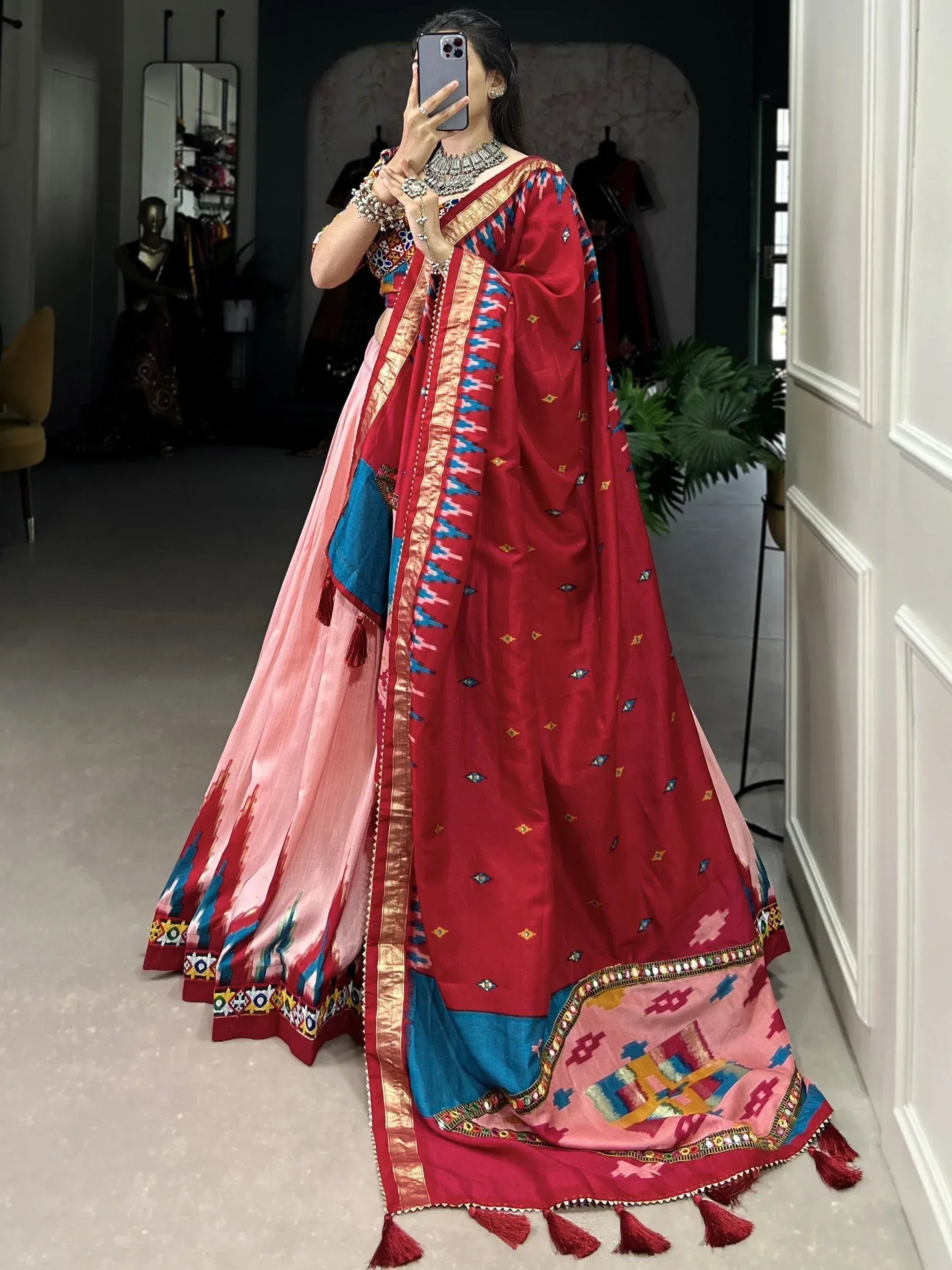 Red and pink ethnic lehenga choli with blue accents, mirror work, and tassels, styled with traditional jewelry