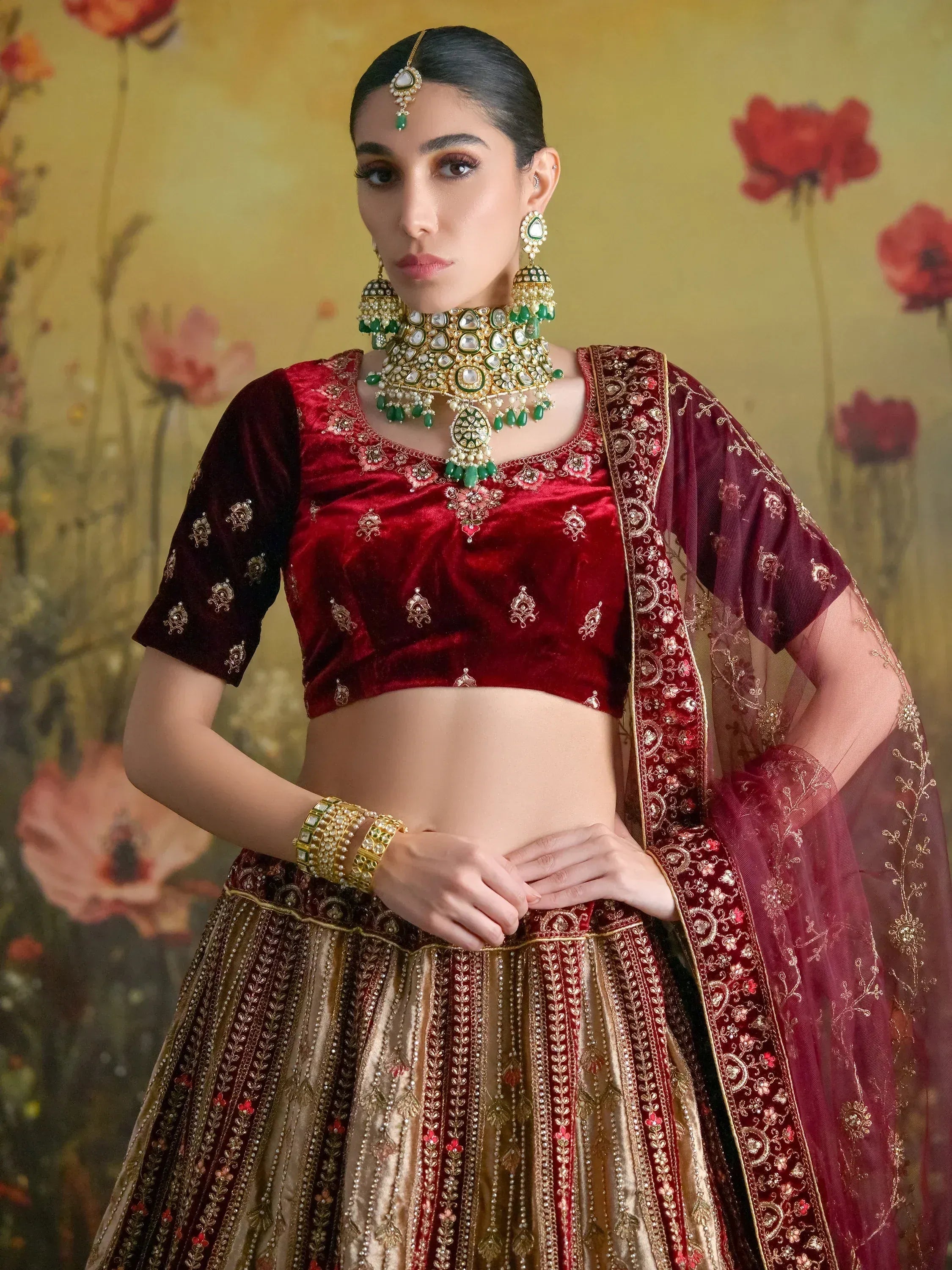 Elegant maroon velvet lehenga choli with gold embroidery, paired with green and gold jewelry for Indian ethnic wear.
