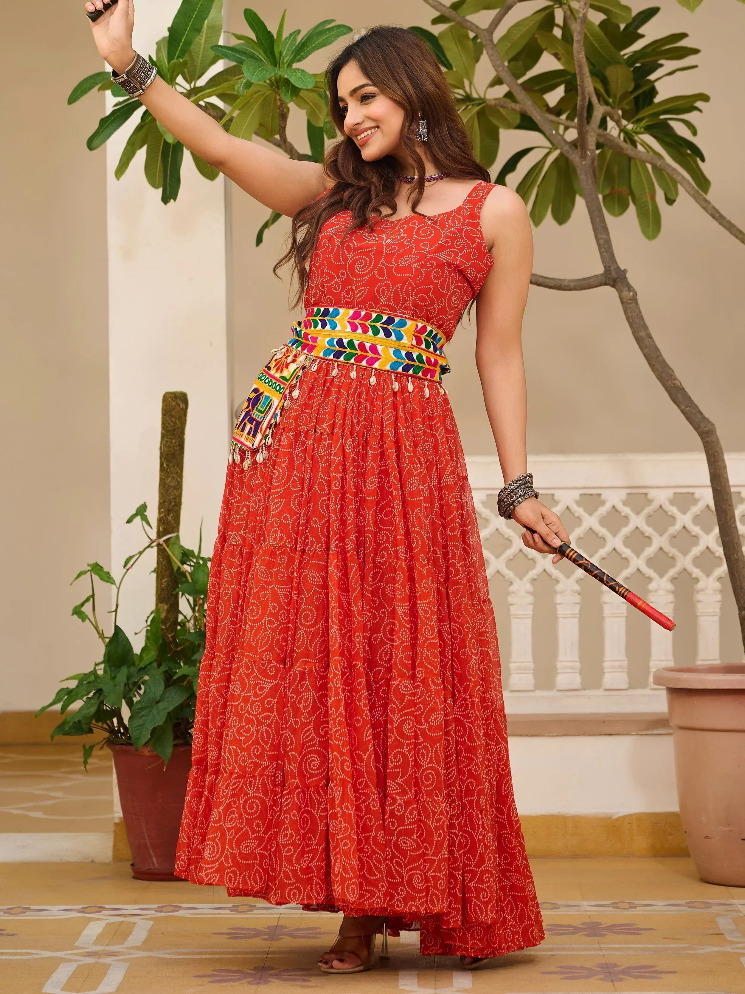 Woman in vibrant red ethnic gown with colorful belt, holding decorative stick, perfect for Indian festive wear.