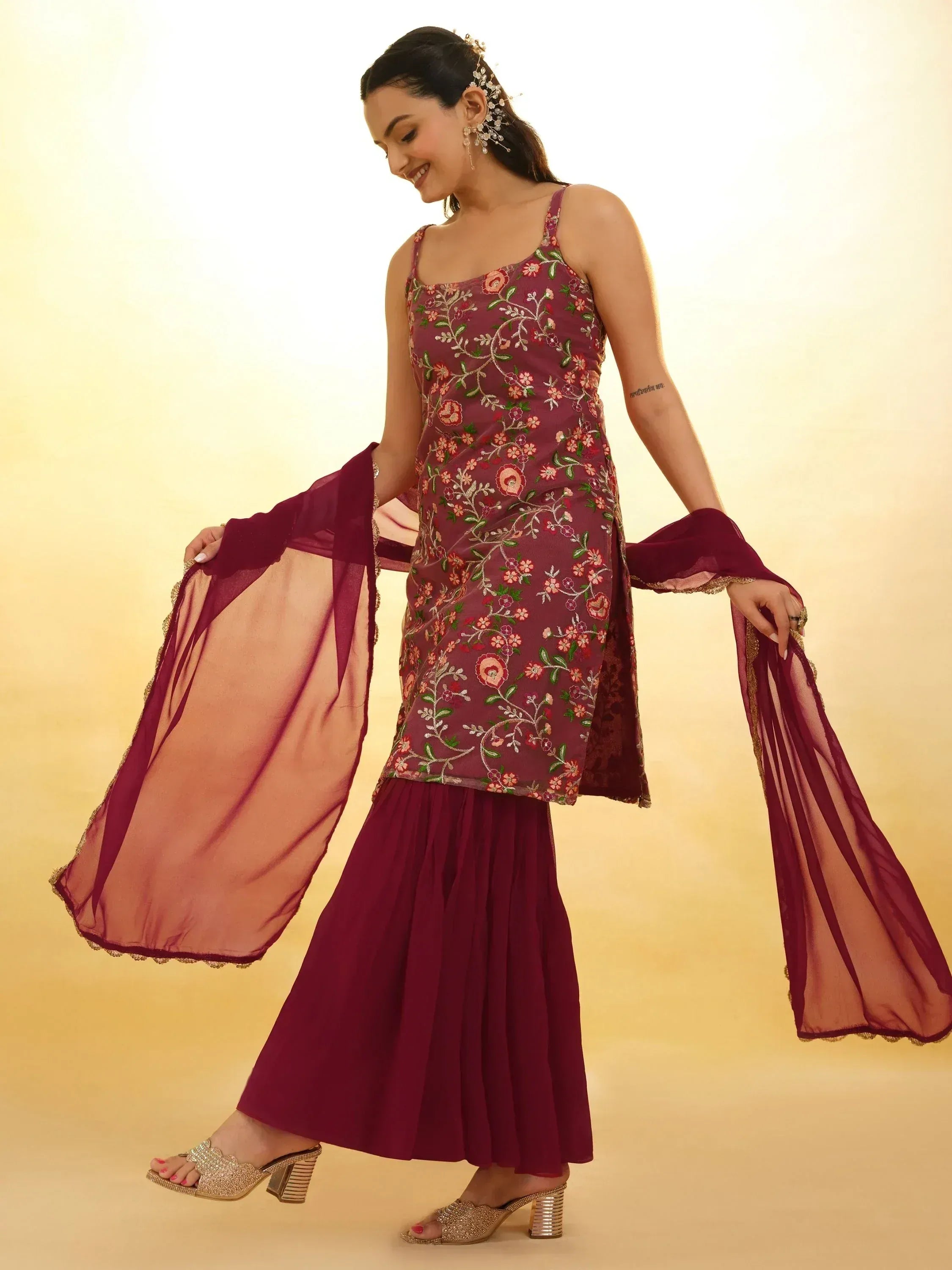 Woman wearing maroon embroidered sharara suit with sheer dupatta and gold heels