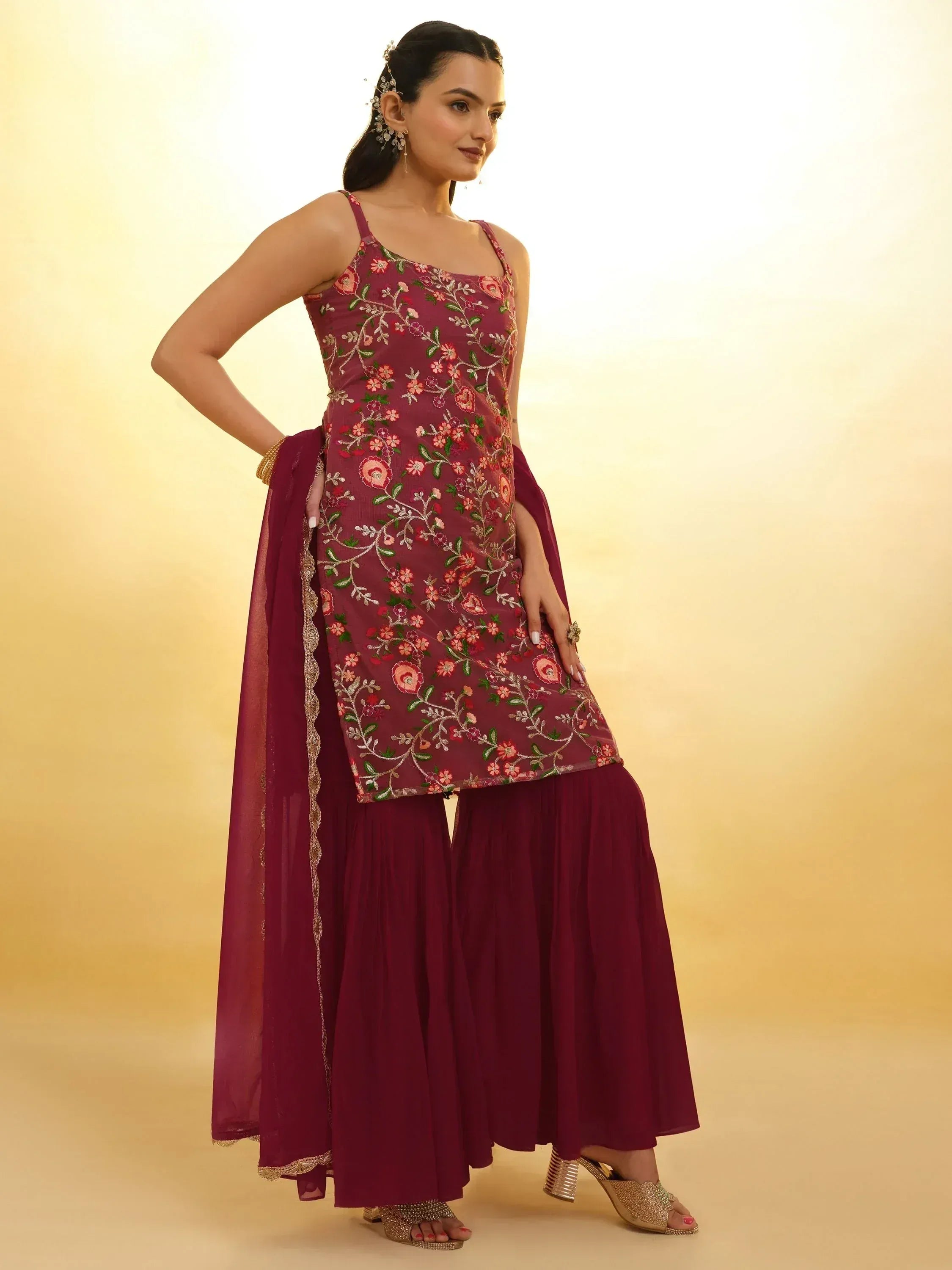 Woman wearing maroon embroidered ethnic sharara suit with dupatta, posing indoors.