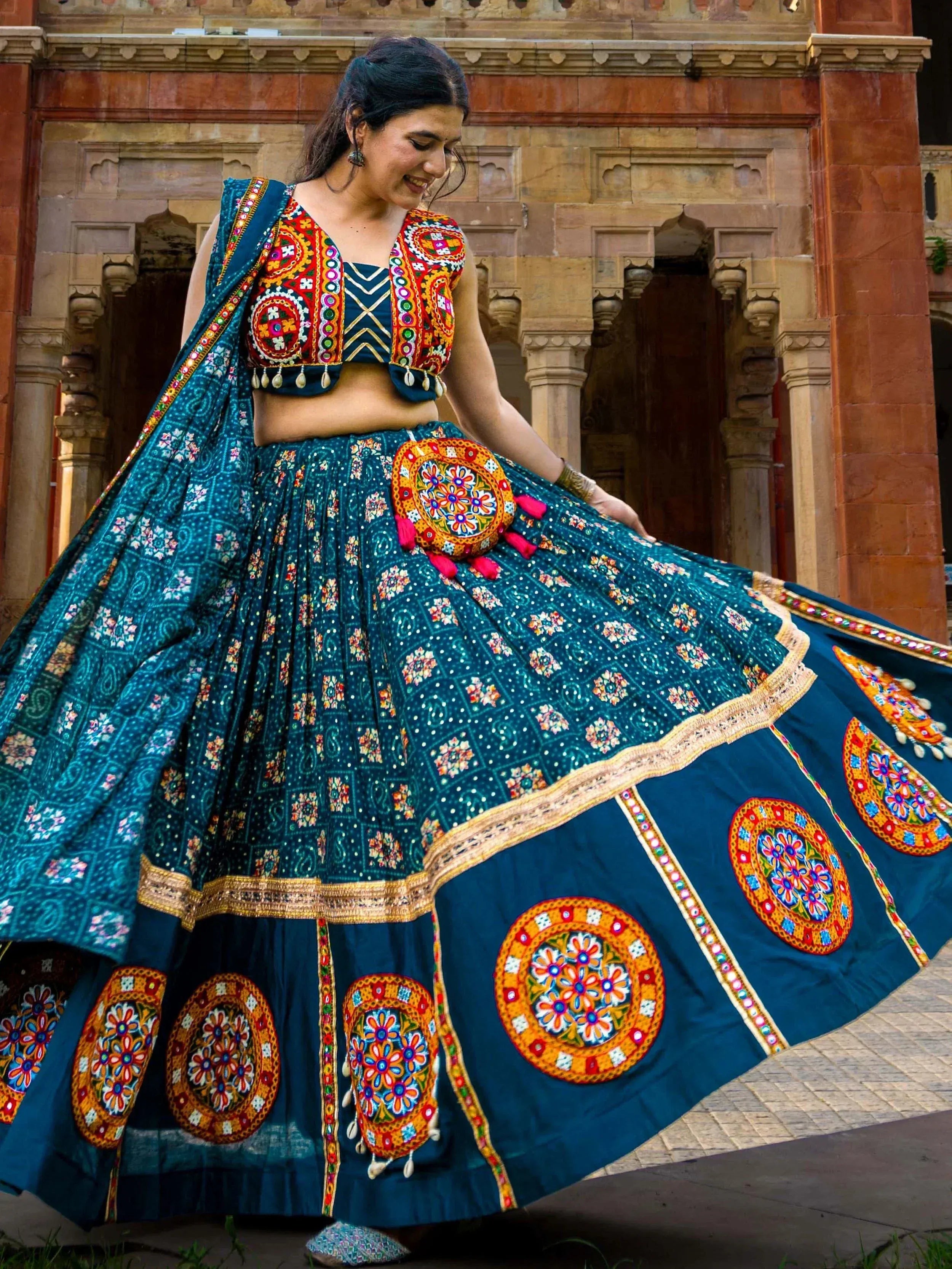 Woman in vibrant teal Indian lehenga choli with mirror work, floral prints, and colorful embroidery, twirling outdoors.