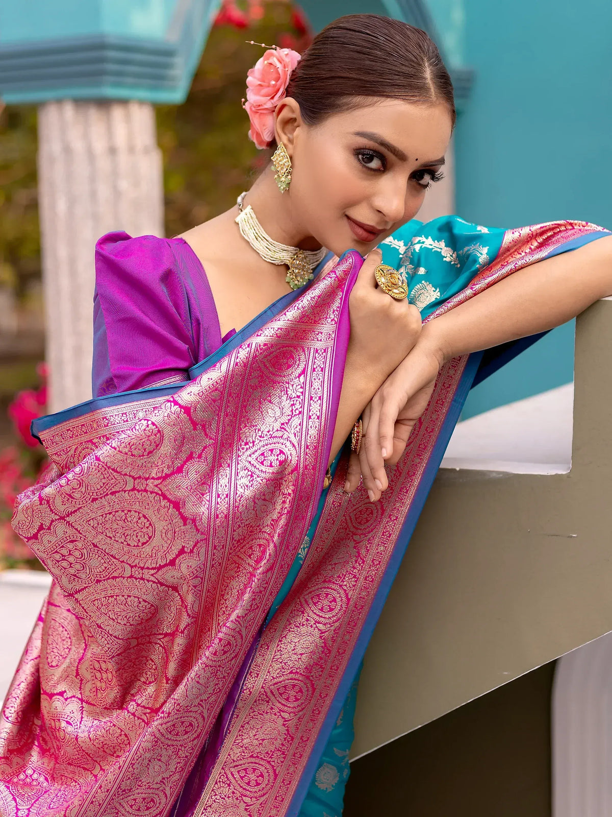 Elegant woman in vibrant pink and blue designer saree with intricate gold paisley patterns, adorned with traditional jewelry.