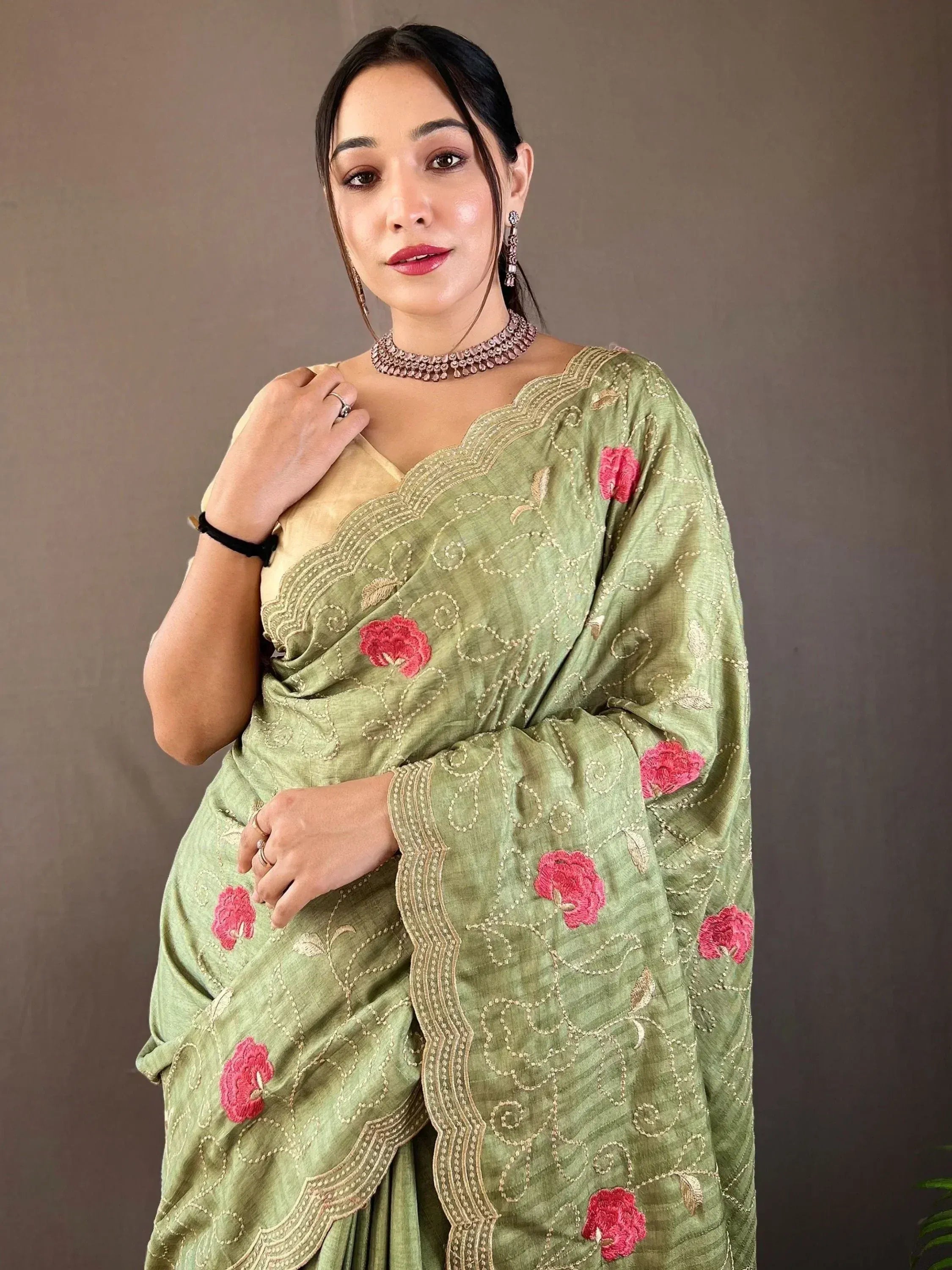 Woman in olive green saree with pink floral embroidery and gold detailing, styled with choker and earrings